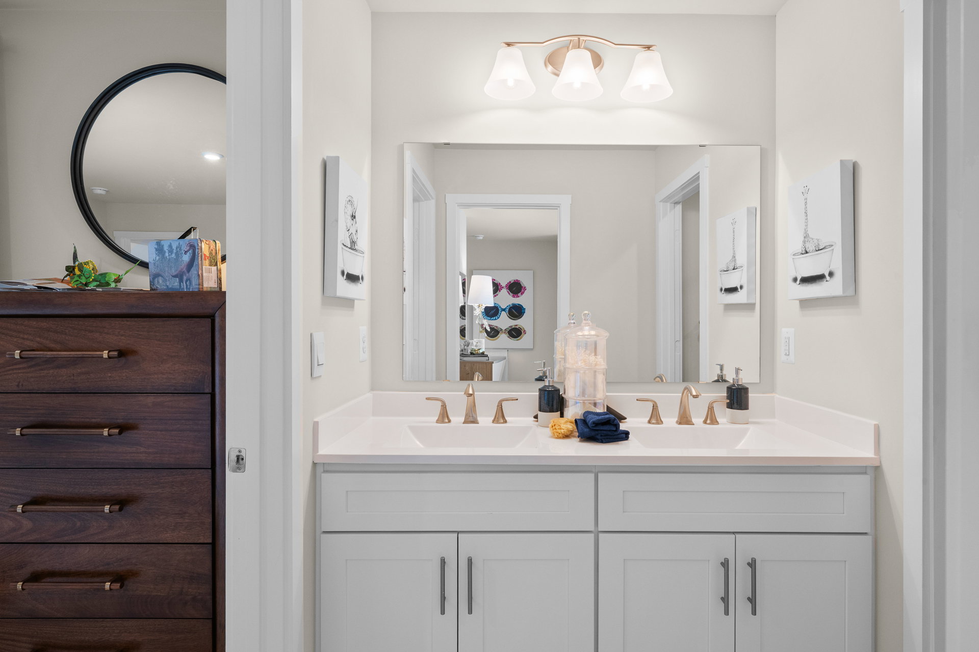 Bright bathroom featuring a double sink vanity with gold fixtures, a round mirror, and a wooden dresser.