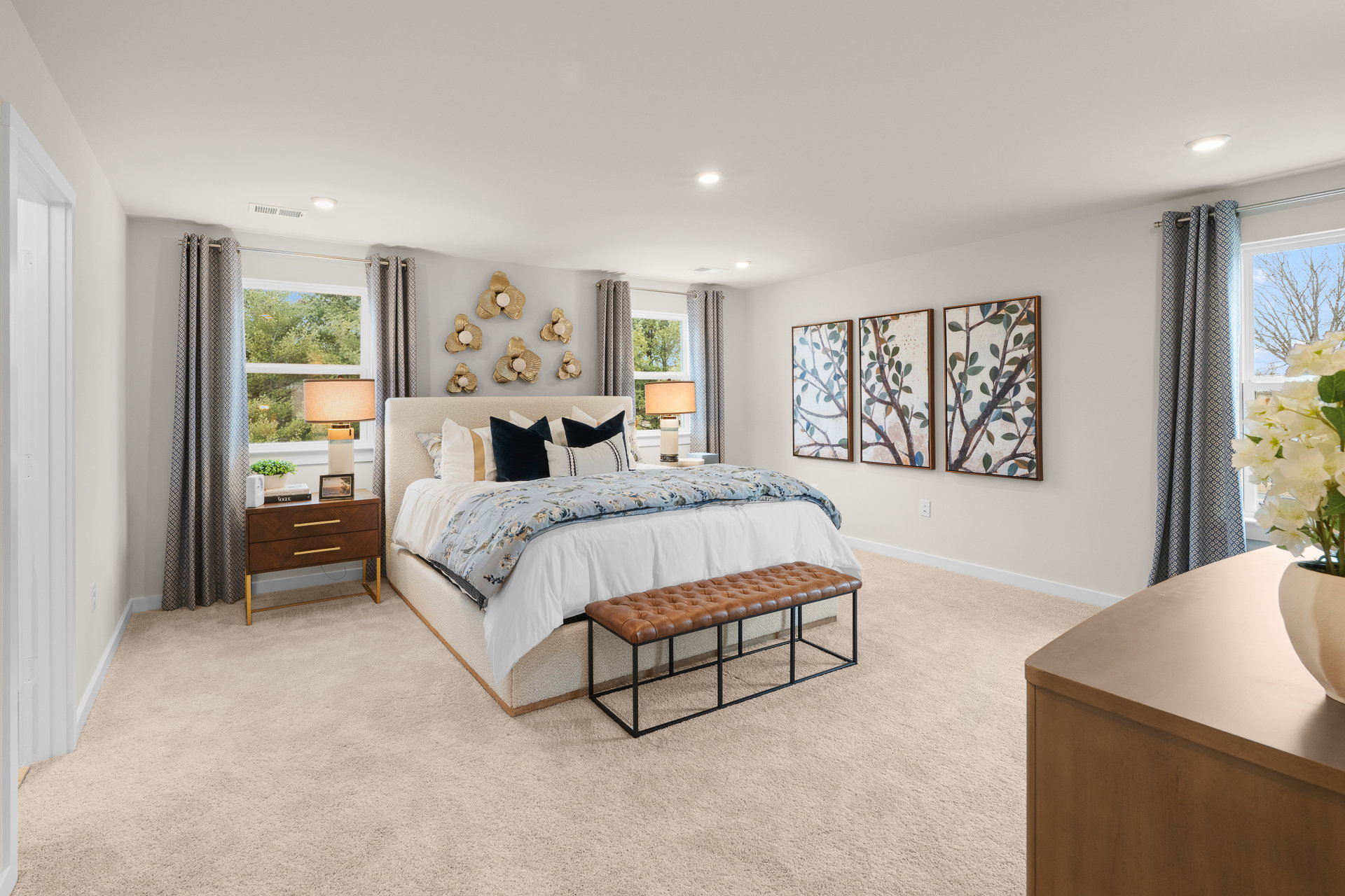 Brightly lit bedroom featuring a large bed with decorative pillows, a bench at the foot, two lamps on side tables, and framed artwork on the walls.