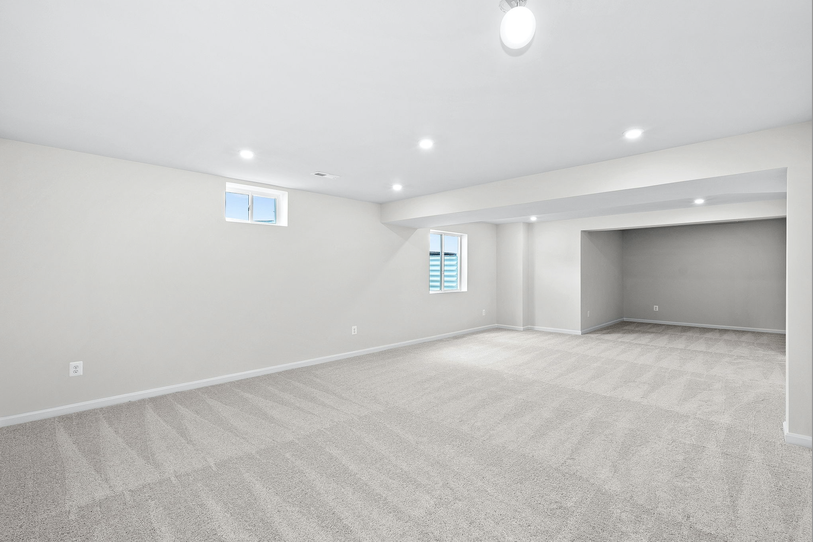 Spacious, modern basement with beige carpet and white walls, featuring recessed lighting and two small windows.