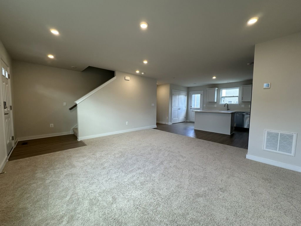 Spacious and bright living room with carpet flooring, modern kitchen, and an inviting entrance showcasing a warm and welcoming atmosphere.