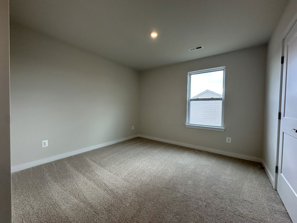 Empty modern bedroom with beige walls, carpeted flooring, and a window allowing natural light.