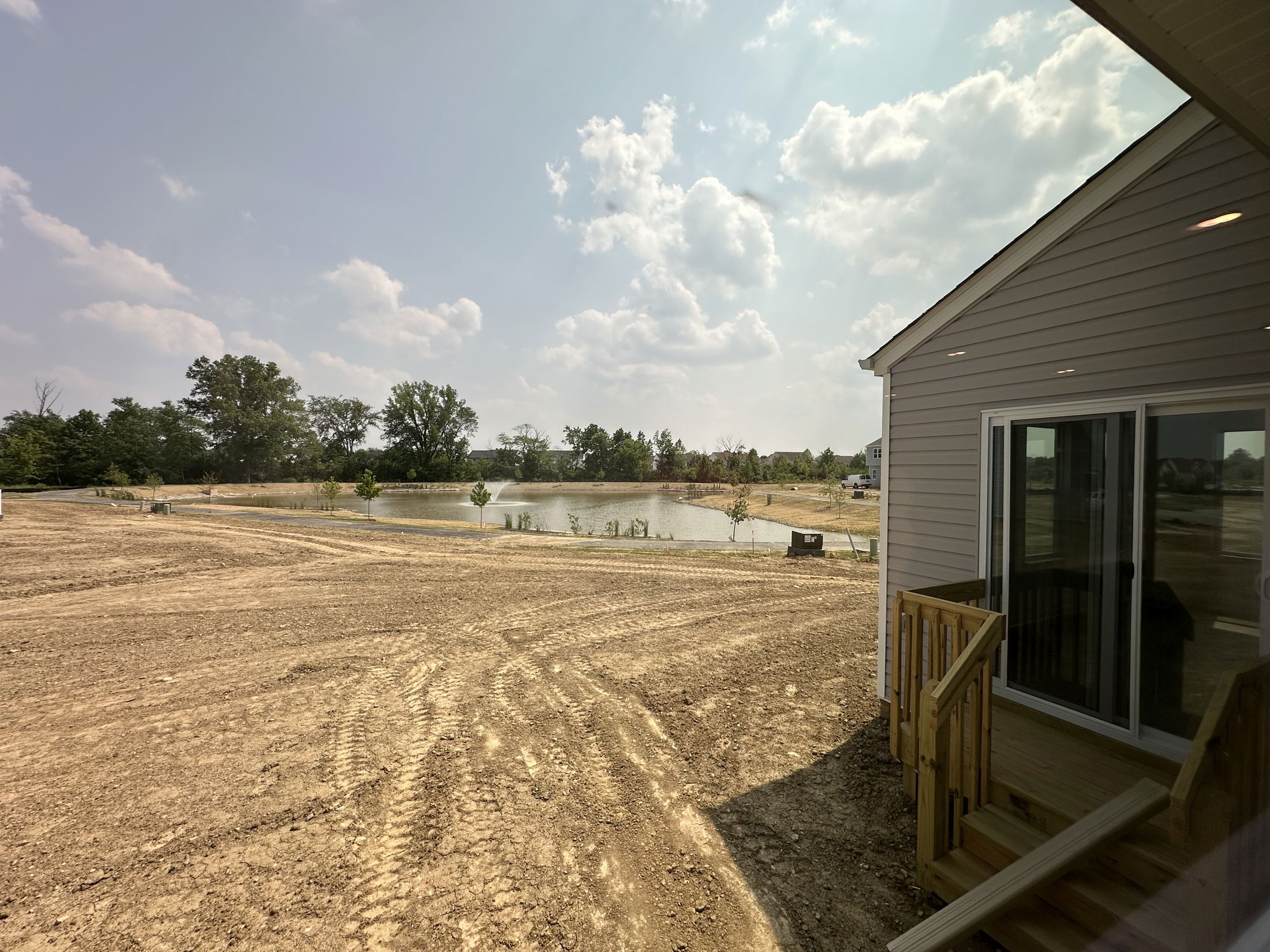 Newly constructed home with a wooden deck overlooking a freshly landscaped yard and small pond under a partly cloudy sky.