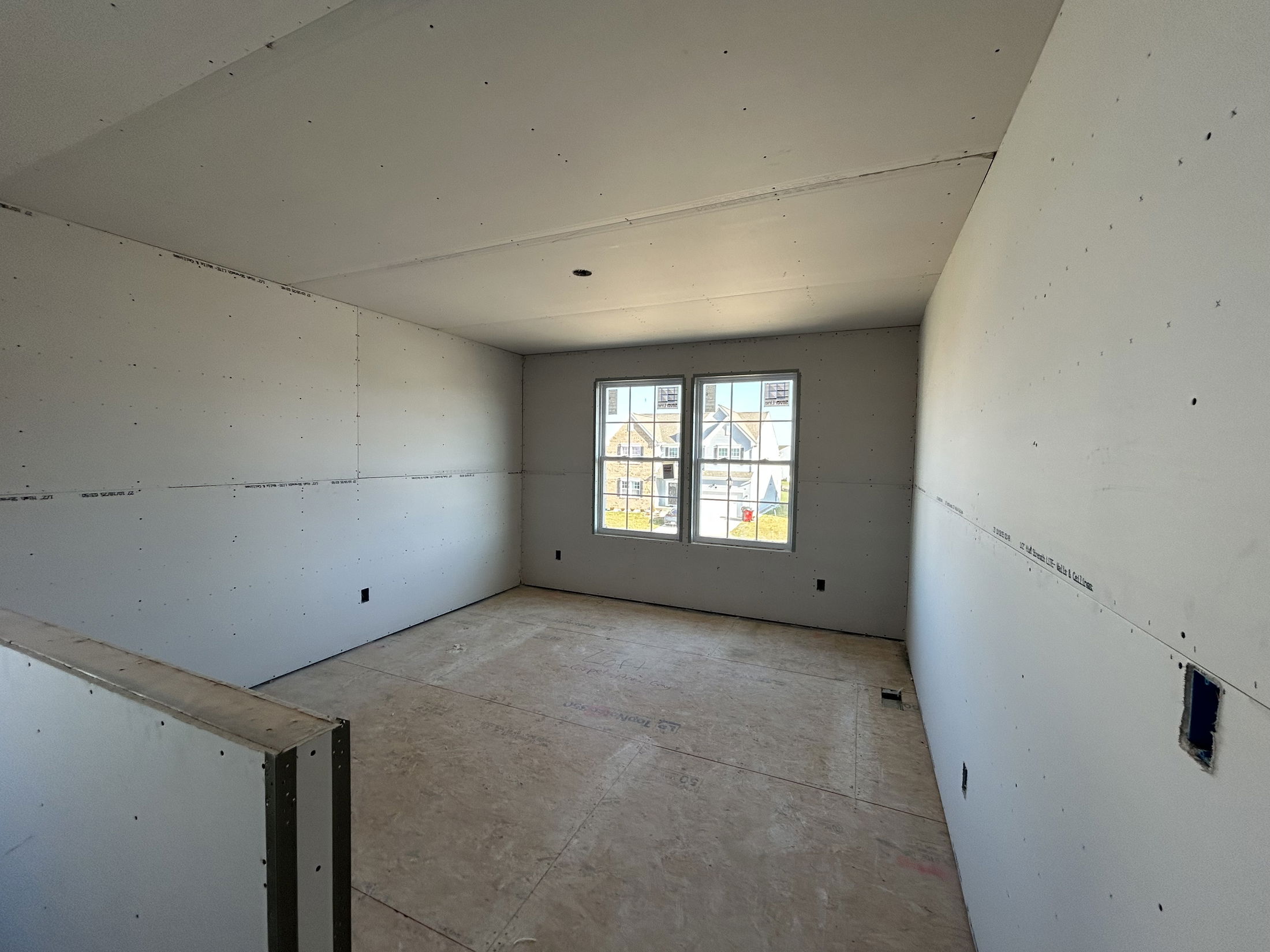 Partially finished room with drywall installation and an unfinished floor, featuring two large windows.