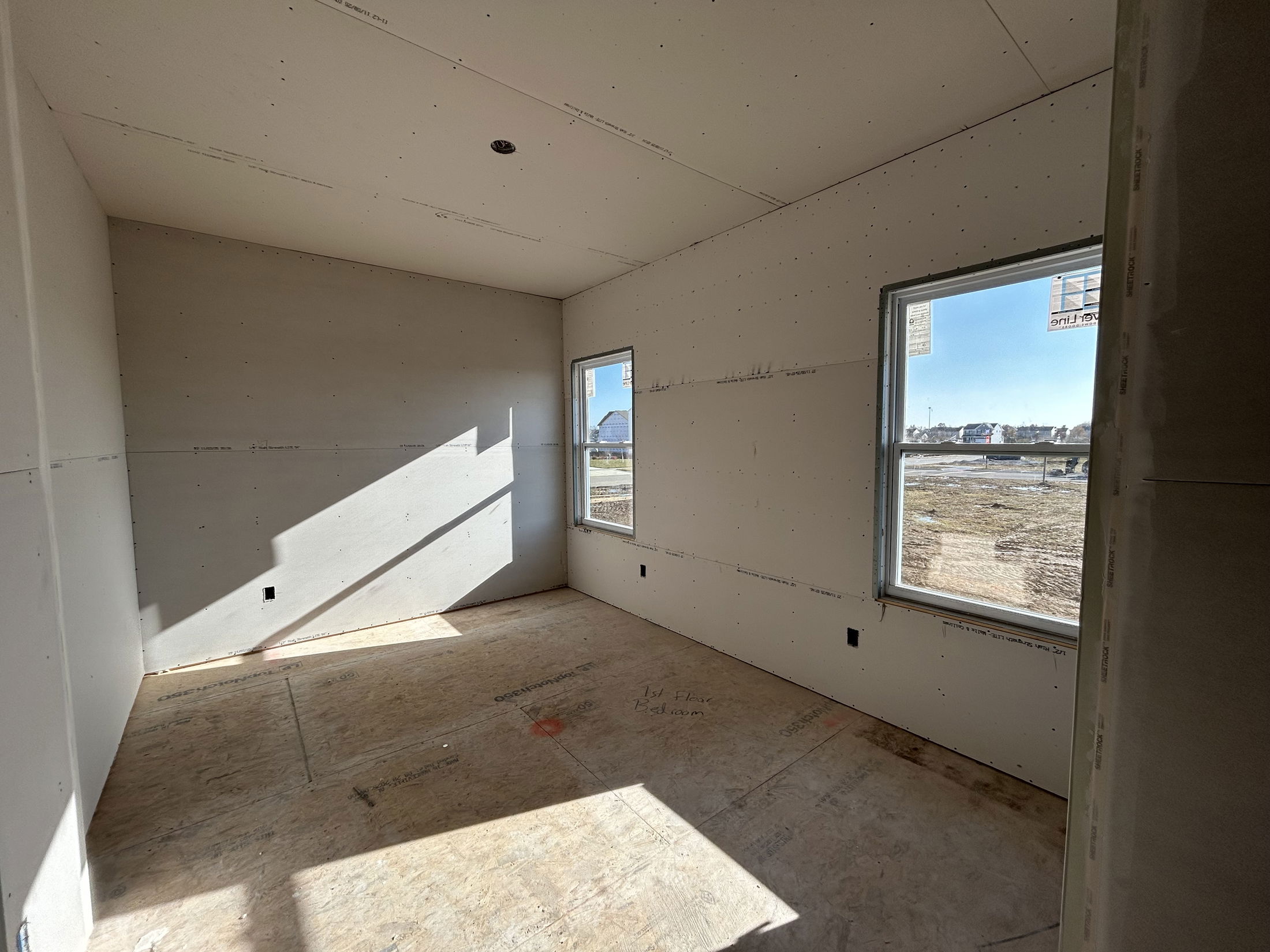Bright, unfinished room with drywall installation and two large windows overlooking a construction site.