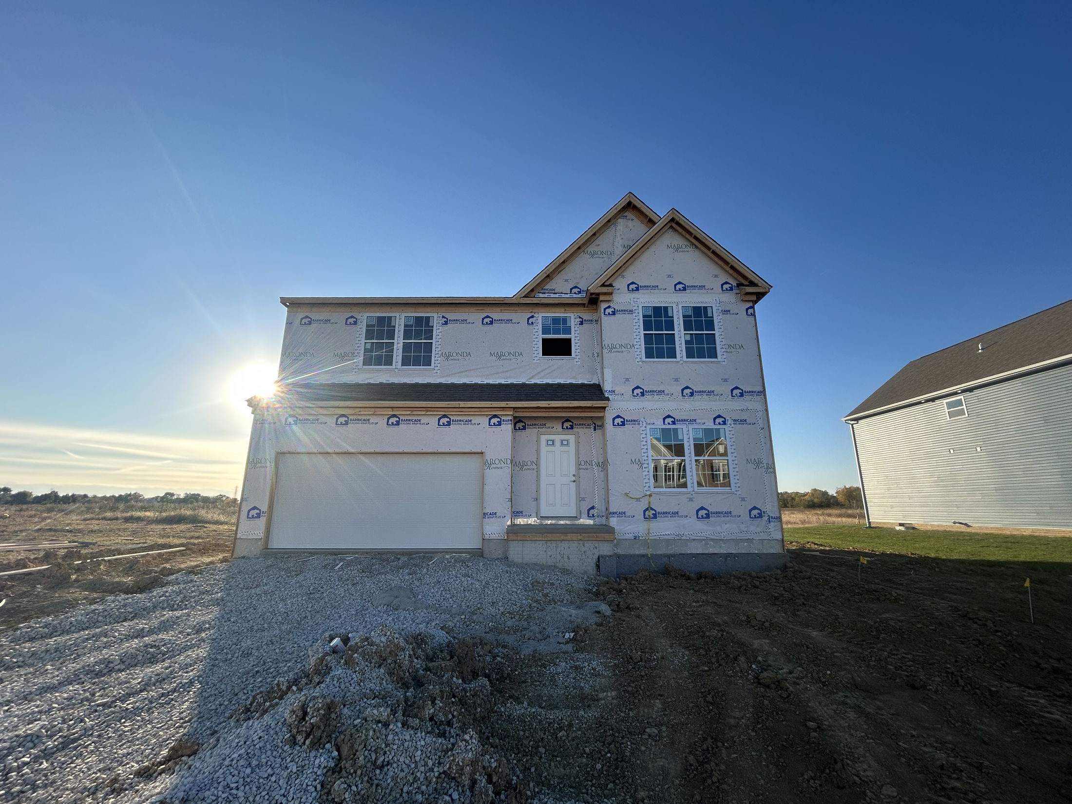 Newly constructed two-story house under clear blue sky with visible wrap and sunlight in a suburban setting.