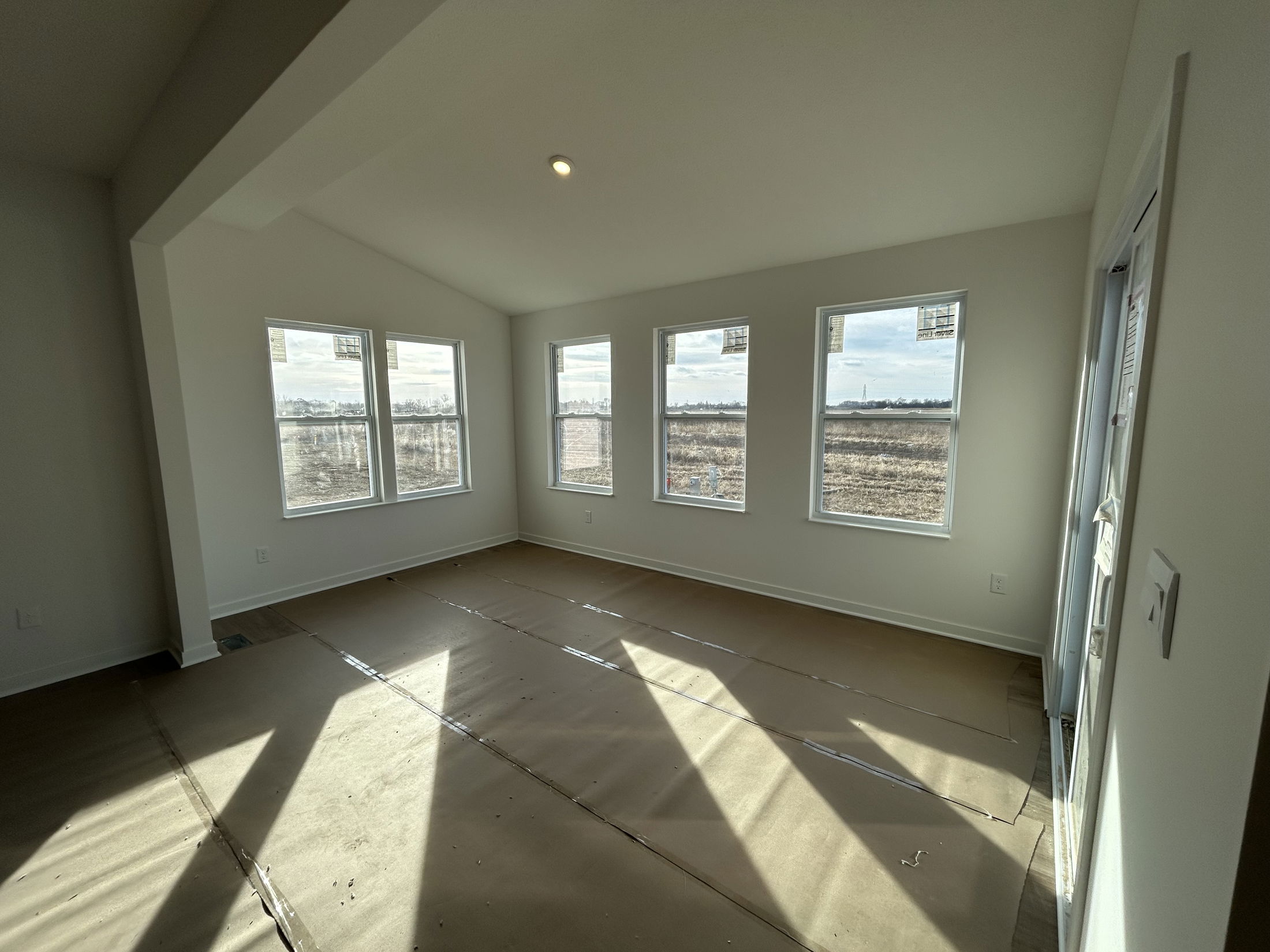 Bright and spacious unfinished living room with large windows overlooking a field, featuring new construction elements and natural light.