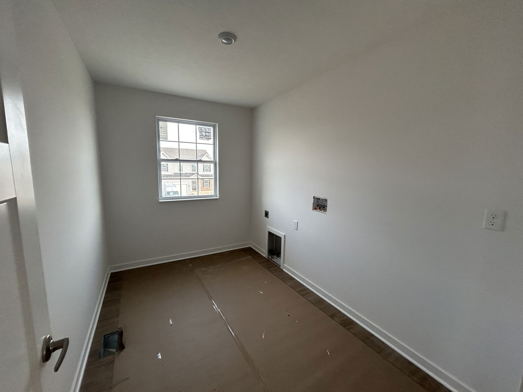 Small, empty room with a single window and light-colored walls, featuring uninstalled electrical outlets and brown floor covering.