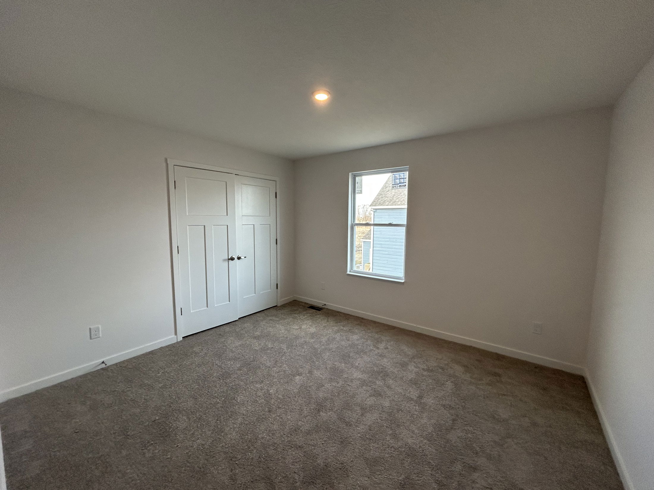 A bright, empty bedroom featuring light gray carpet, white walls, and a double closet with a window allowing natural light.