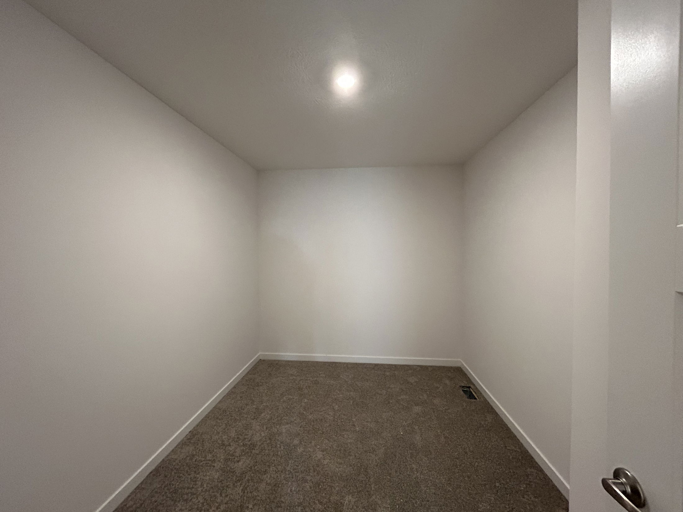 Interior view of an empty room with white walls and a carpeted floor, featuring a ceiling light and an adjacent door.