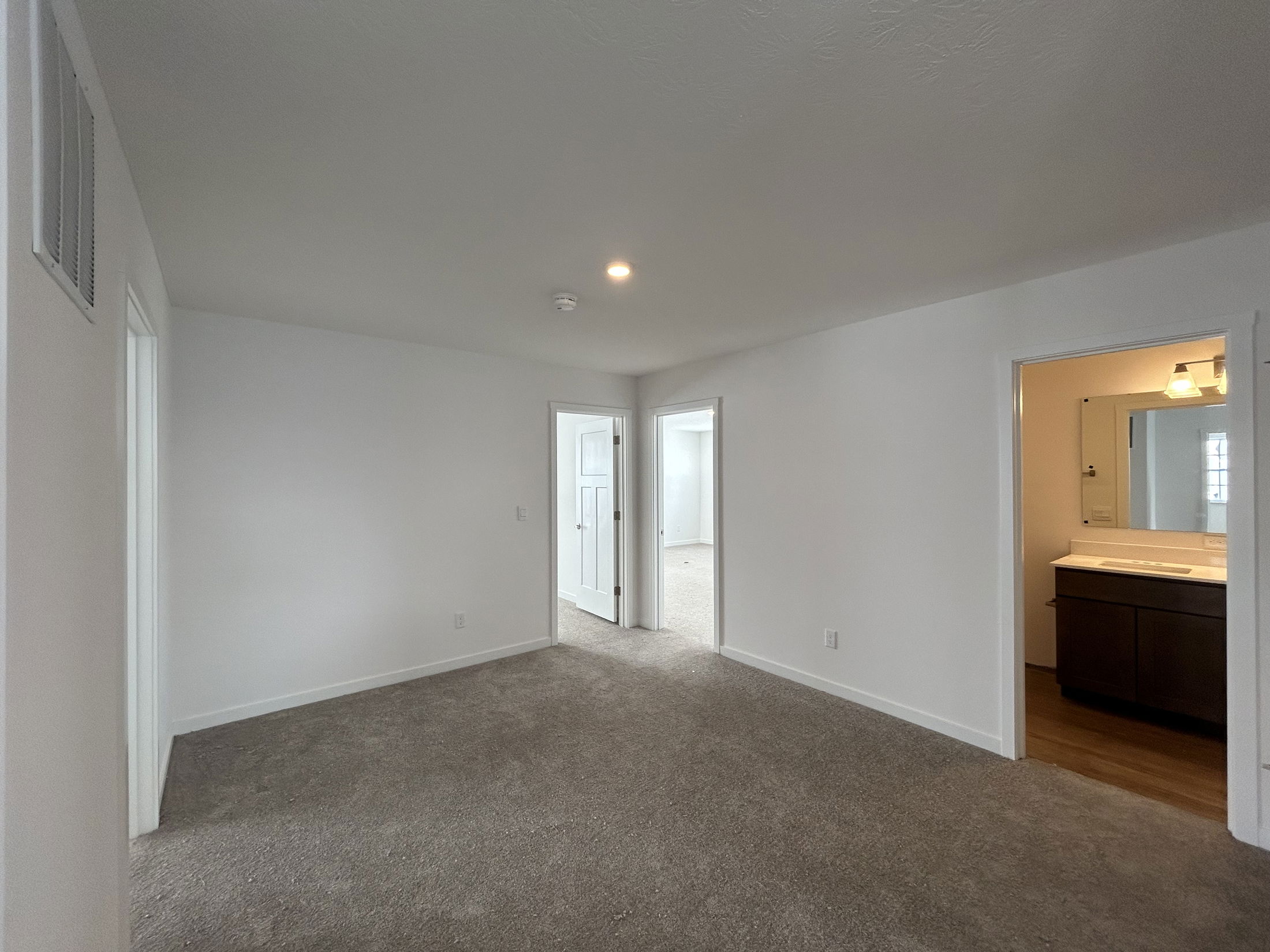Spacious and well-lit interior of a modern apartment featuring carpeted flooring, open doorways, and a view of a bathroom with vanity.