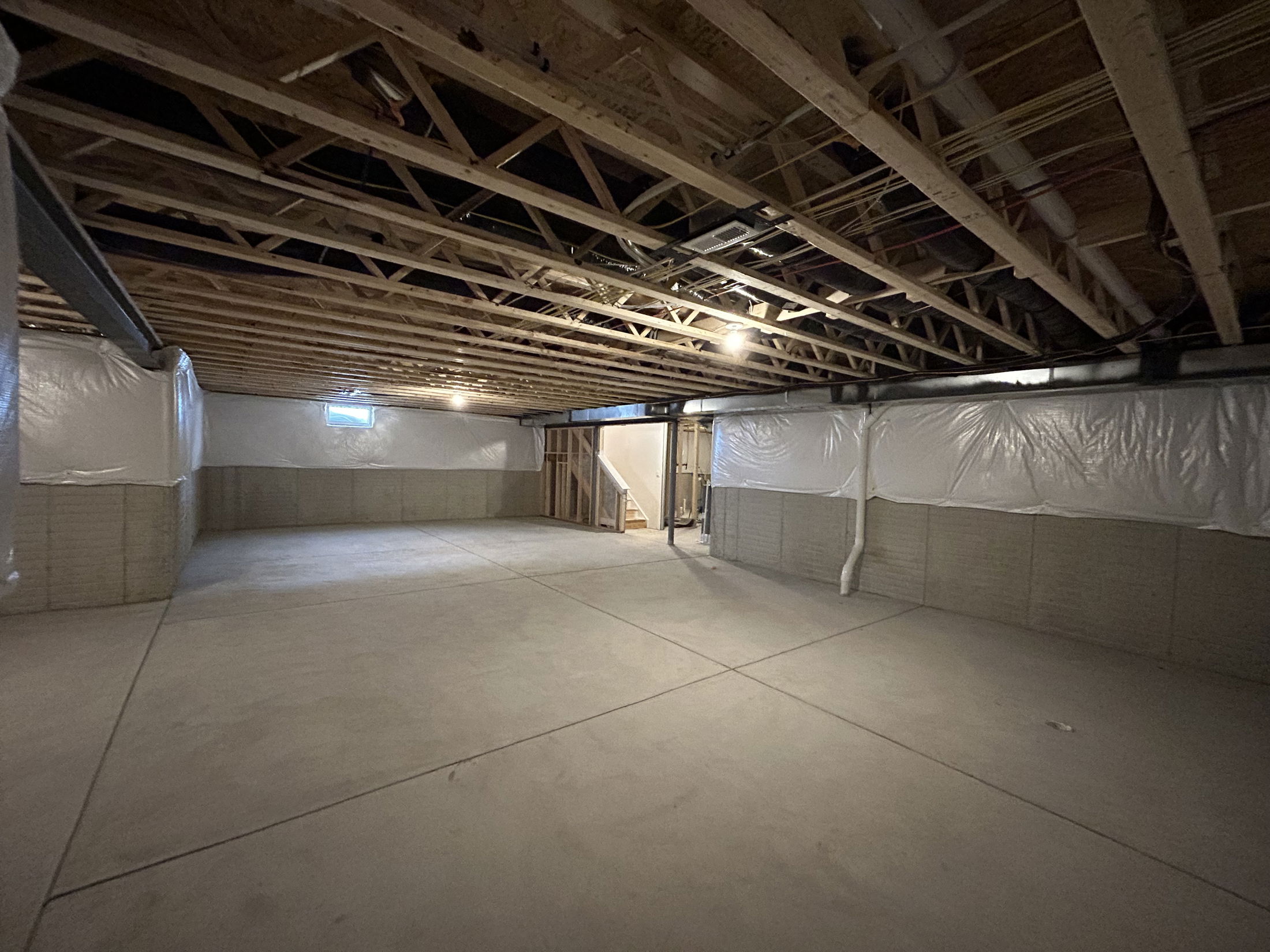Spacious unfinished basement with exposed wooden beams, concrete flooring, and plastic-wrapped walls.