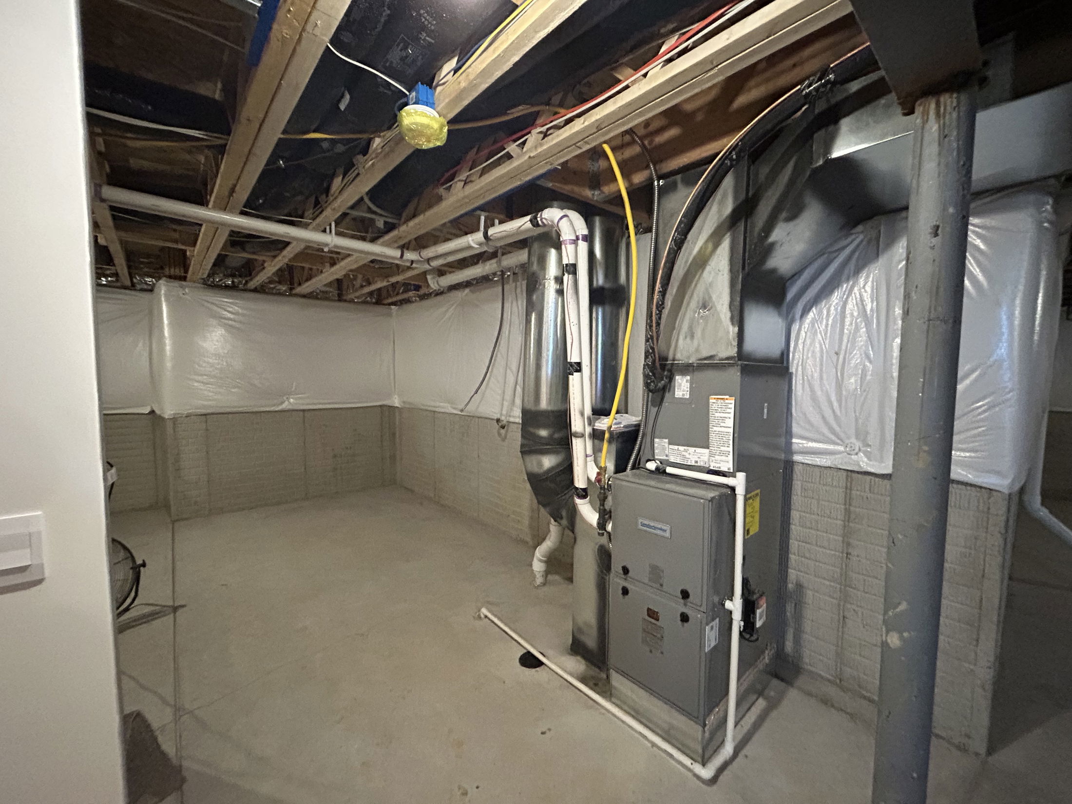 A clean and unfinished basement featuring exposed beams, a furnace, and plastic-wrapped walls.