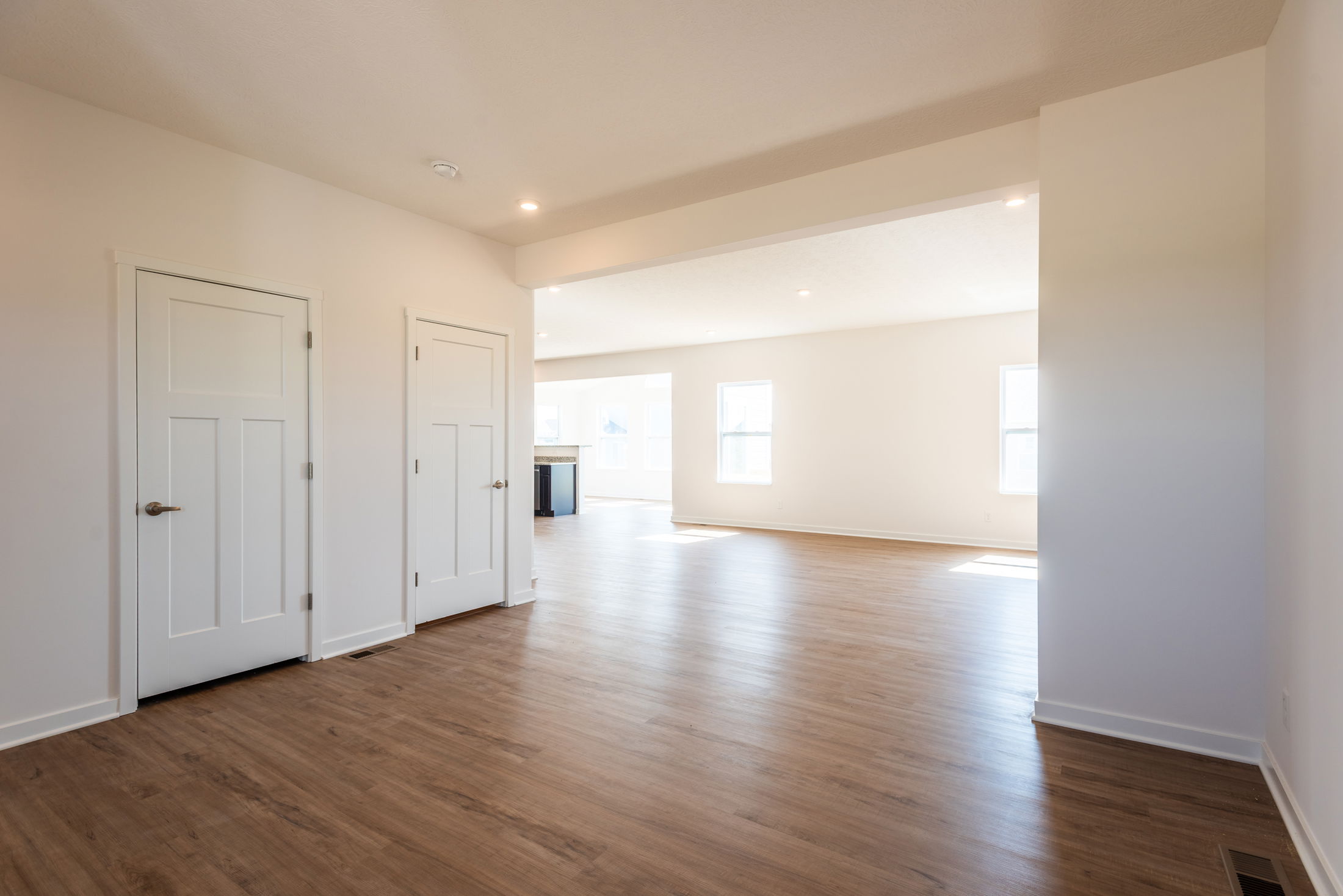 Bright, spacious empty room with hardwood floors and white walls, featuring two closed doors and natural light pouring through multiple windows.