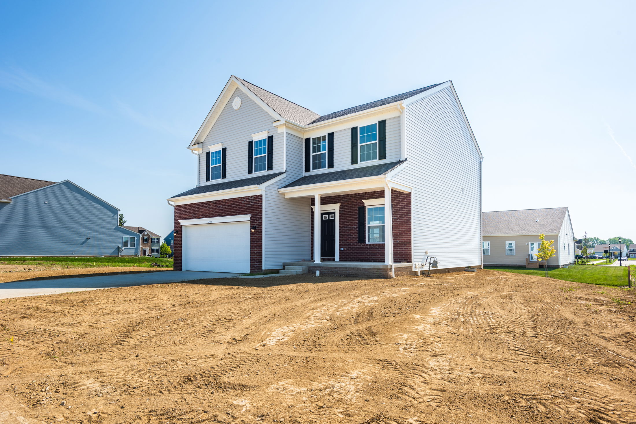 Two-story suburban house with brick and siding exterior, featuring a garage and bare yard under a clear blue sky.