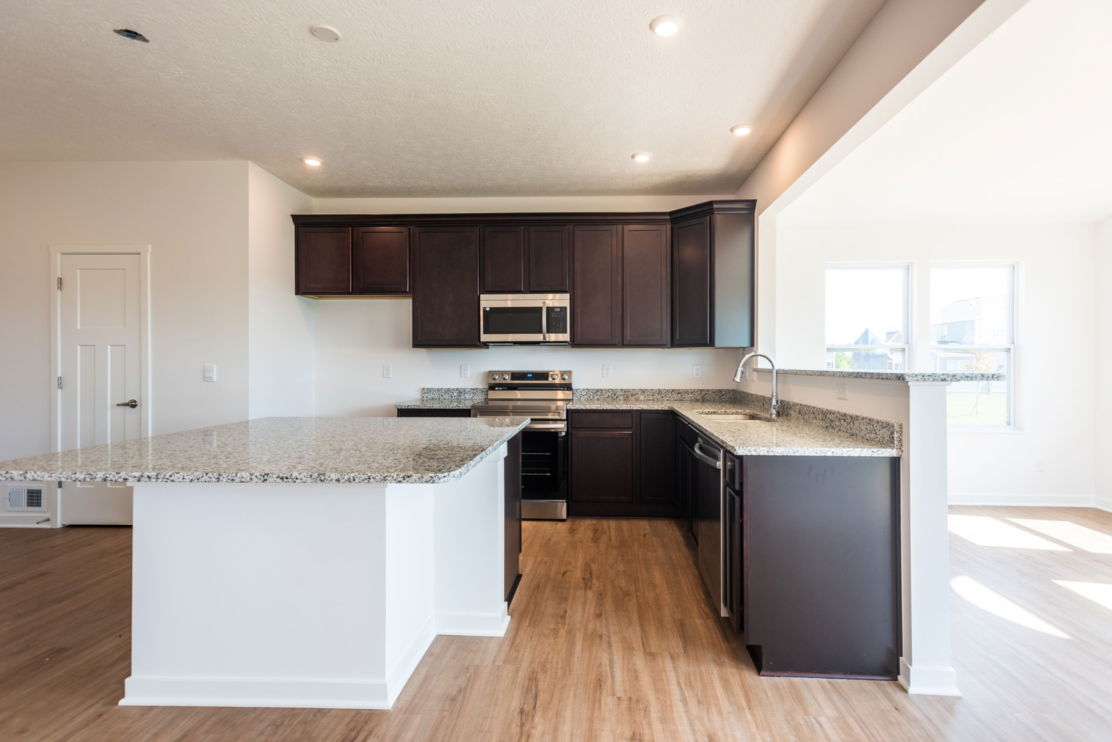 Modern kitchen with dark wood cabinets, a large granite island, stainless steel appliances, and wooden flooring.