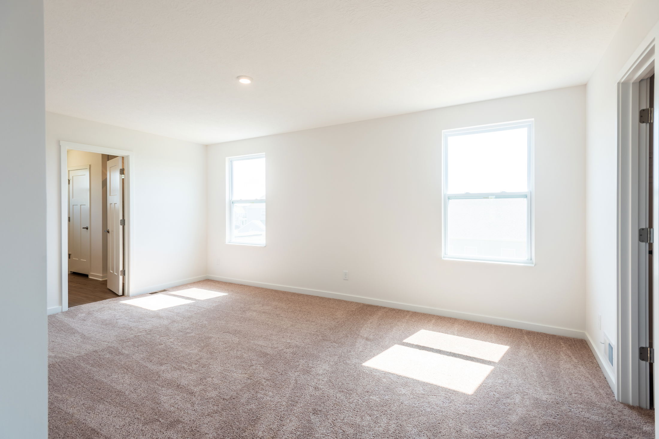 Spacious, well-lit bedroom with beige carpet and two windows, connected to a hallway.