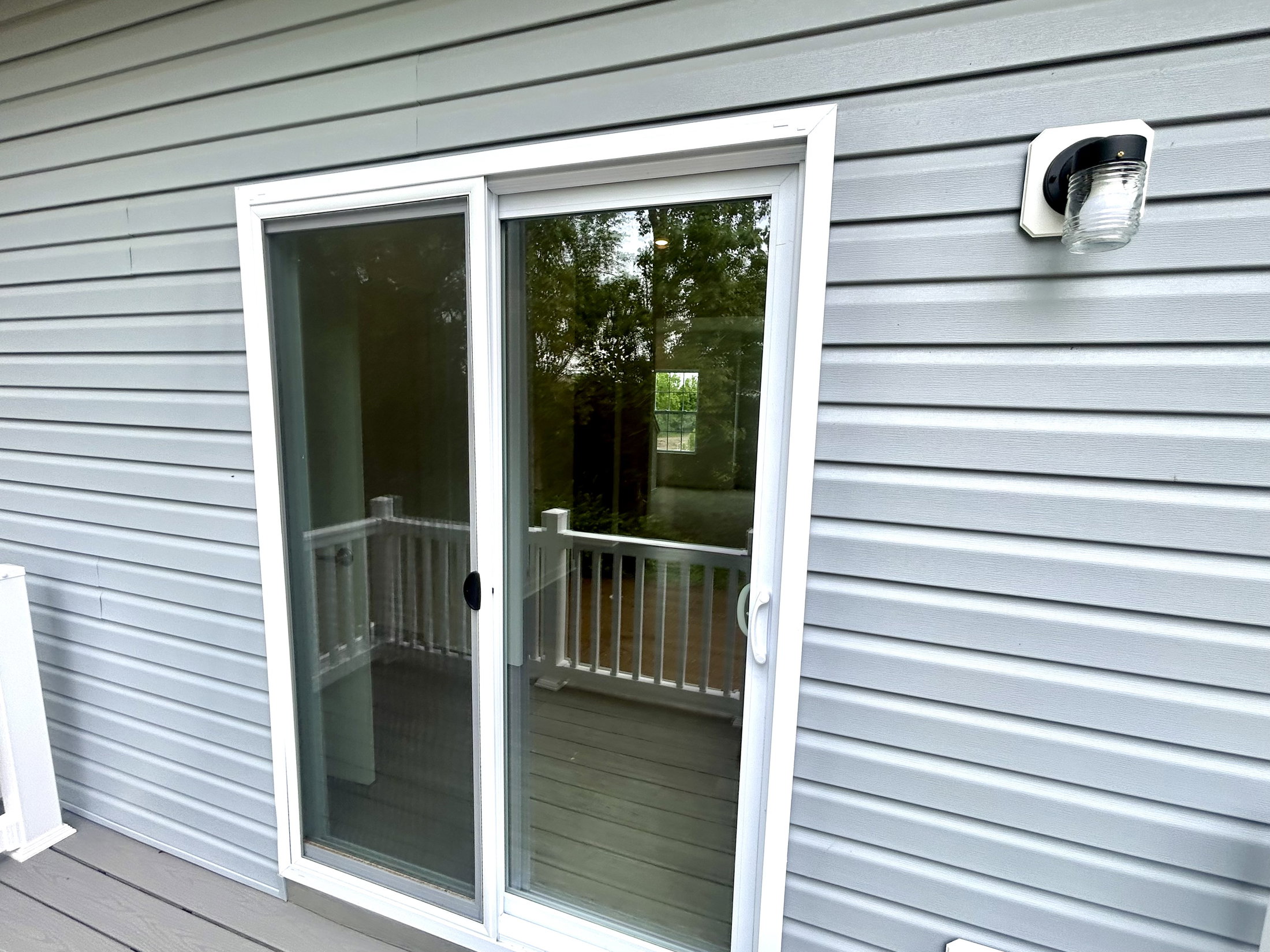 Sliding glass door with a view of a balcony, framed by gray siding and an outdoor light fixture.