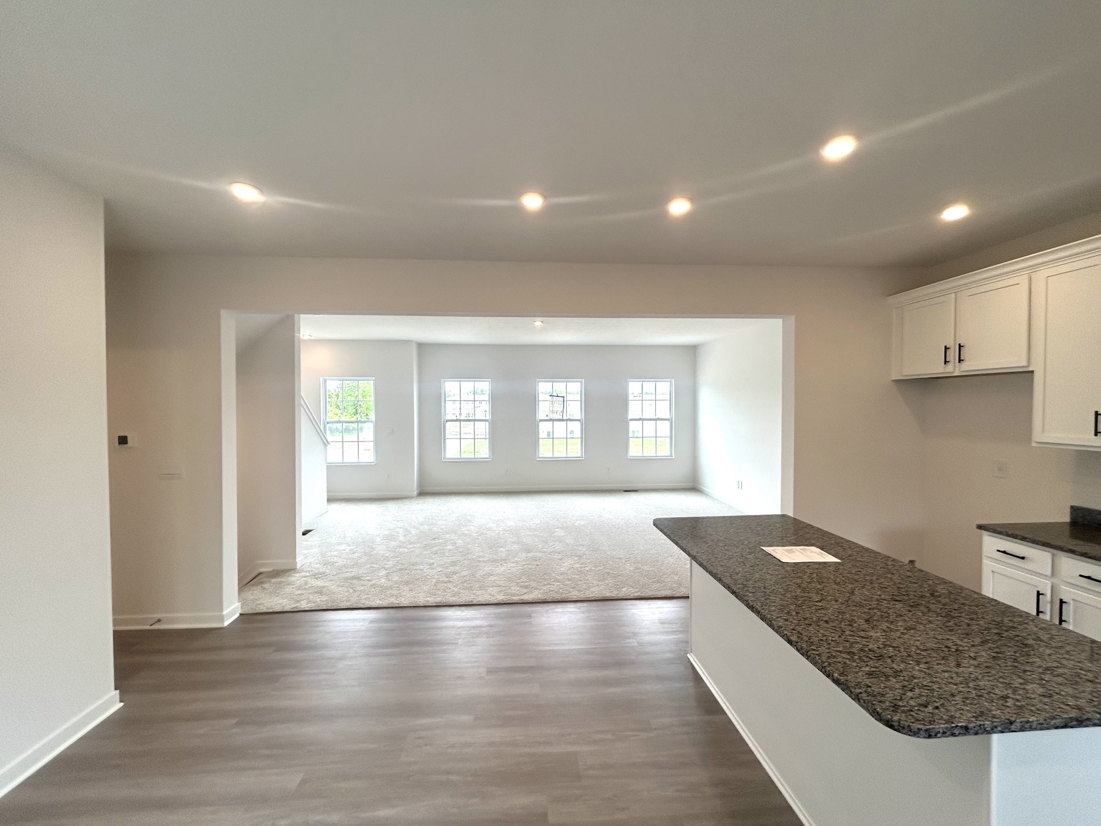 Spacious modern kitchen view with granite countertops leading into a bright living area showcasing large windows and soft carpeting.