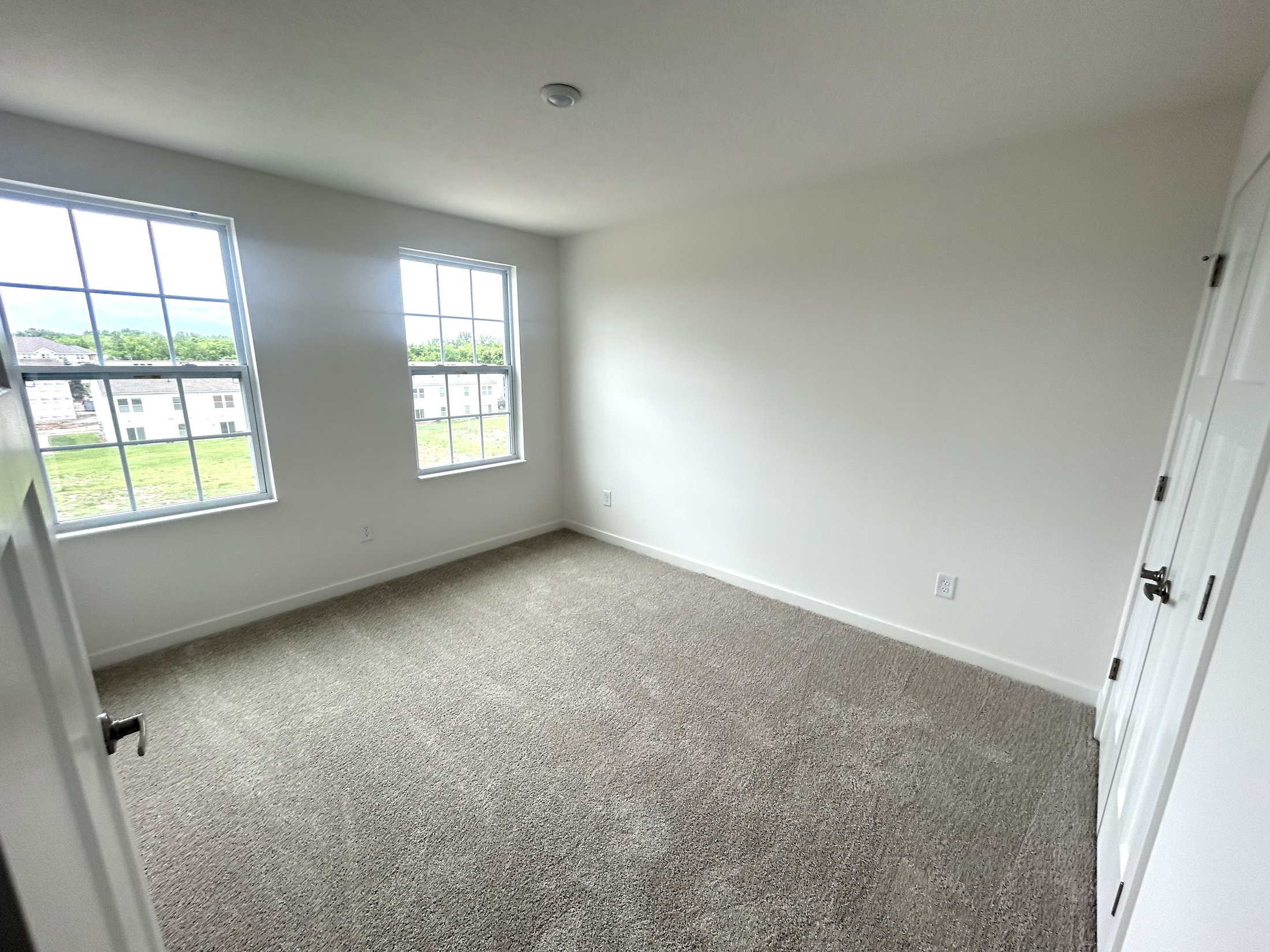 Bright and spacious empty bedroom with two large windows and carpeted flooring.
