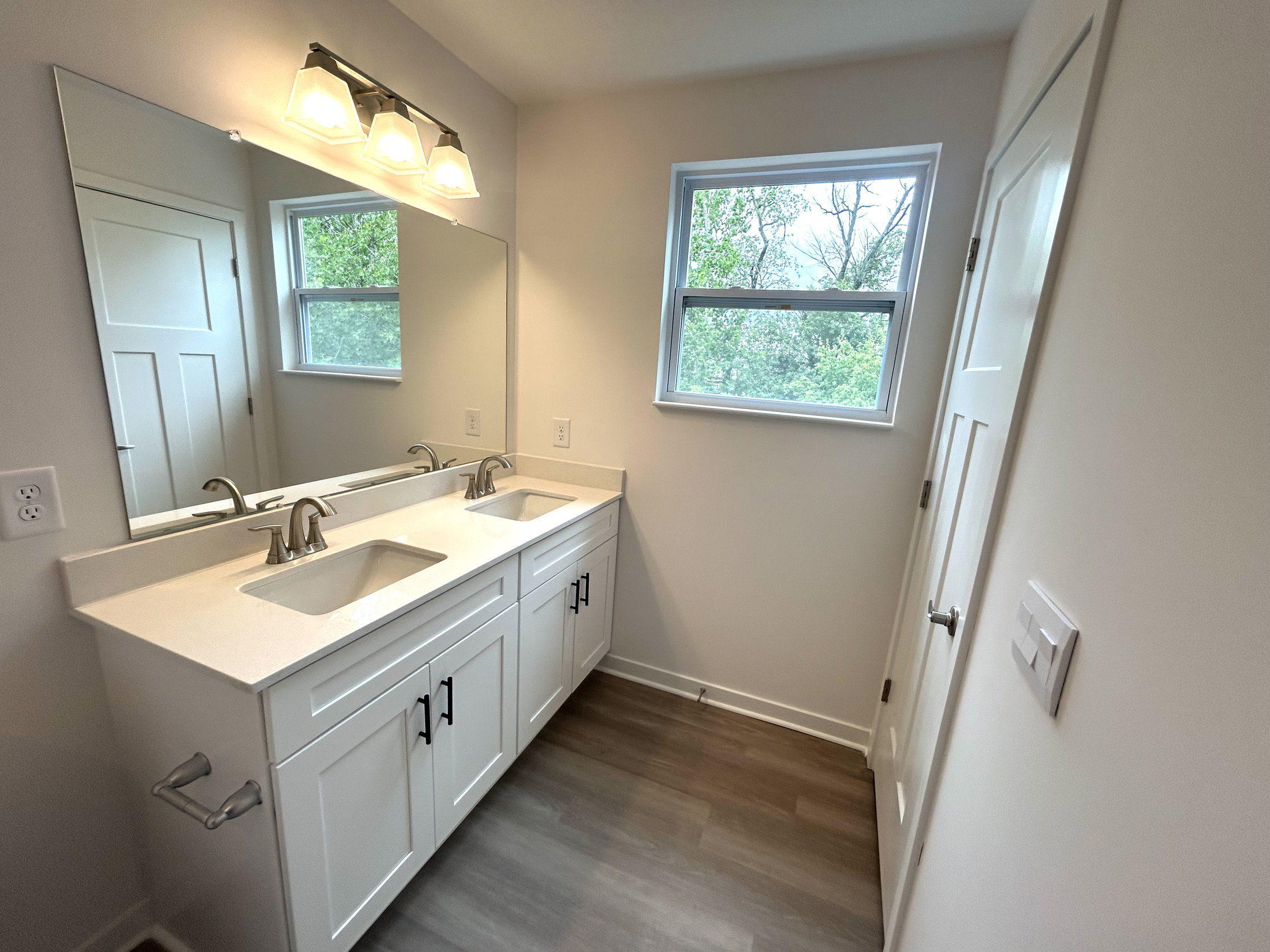 Modern bathroom interior featuring double sinks, a large mirror, and natural light from a nearby window.