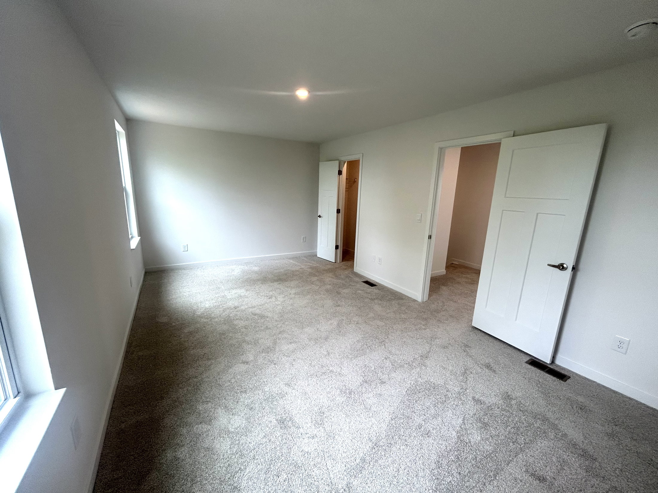 Spacious and bright empty room with carpeted flooring and white walls, featuring multiple doors and large windows for natural light.