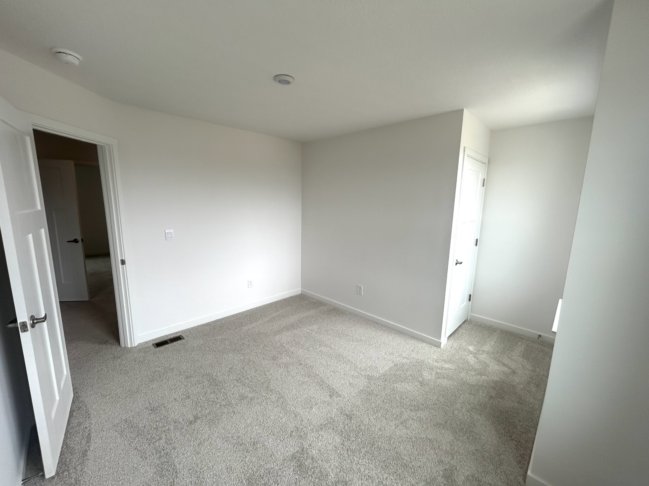 Spacious, empty bedroom with beige carpet and white walls, featuring an open doorway and a bright atmosphere.