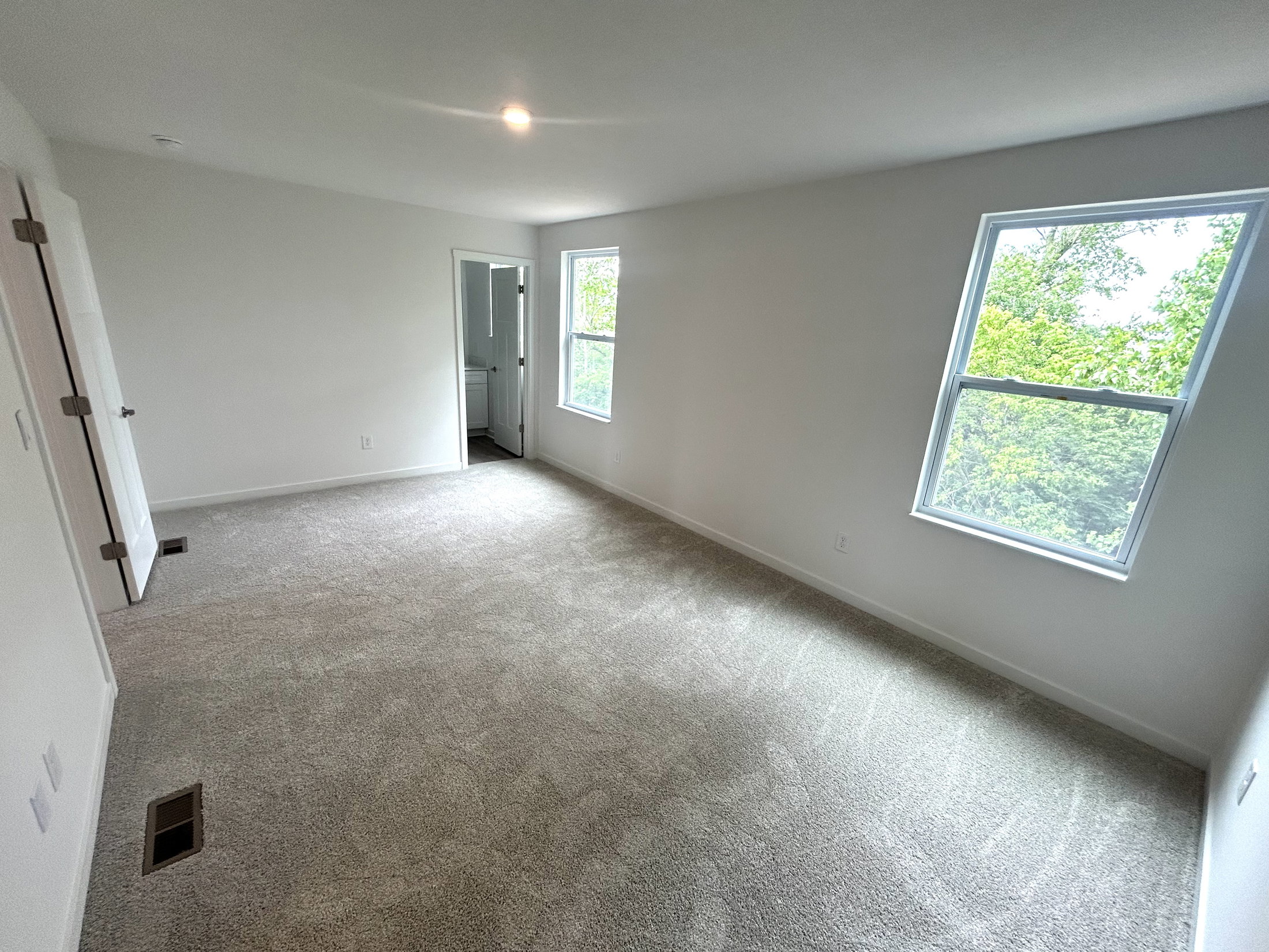 Spacious, empty bedroom with light gray carpet and large windows overlooking greenery.