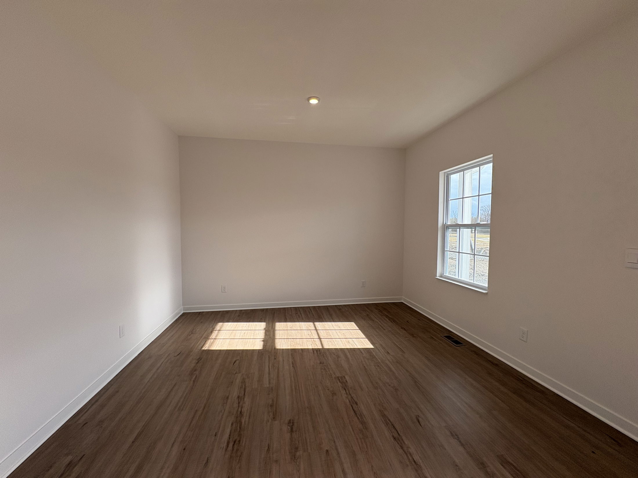 Empty living room with natural light streaming through a window onto the hardwood floor and white walls.