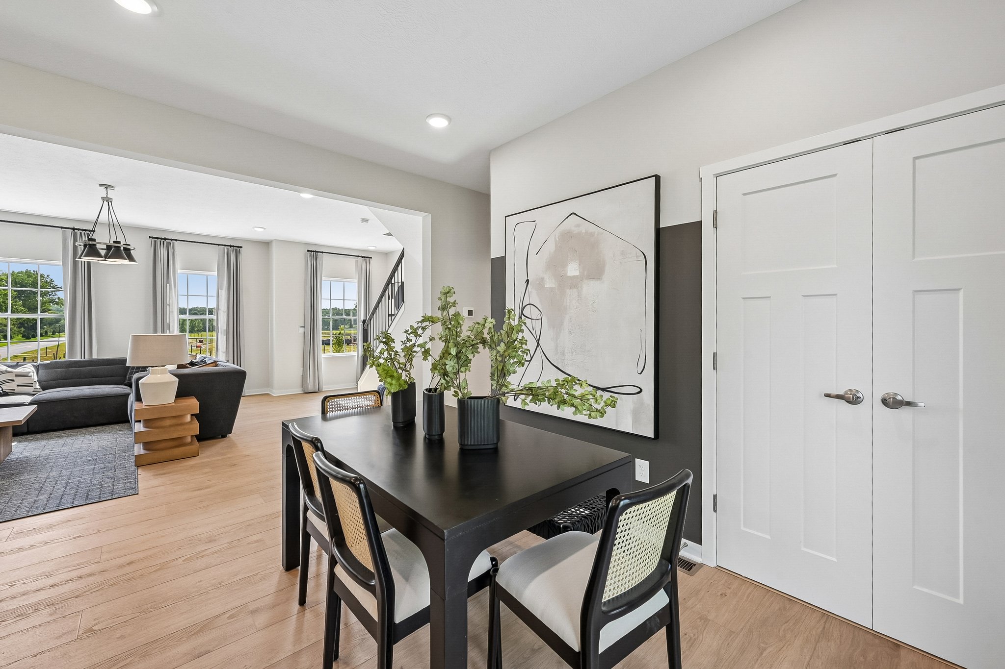 Dining area with a black table, chairs, and a vase of greenery, leading to a bright living room with large windows.