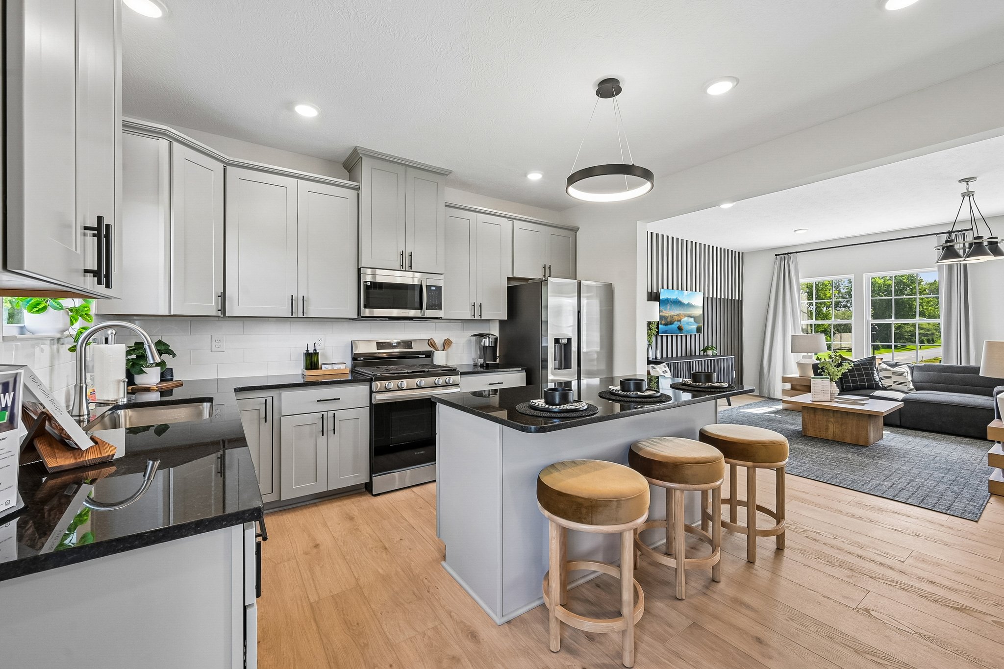 Modern kitchen with gray cabinetry, stainless steel appliances, a central island with bar stools, and a living room visible in the background.