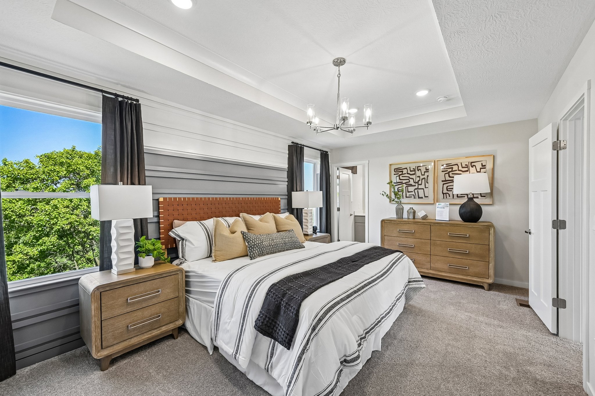 A modern bedroom featuring a large bed with decorative pillows, nightstands with lamps, a window overlooking trees, and a wooden dresser.