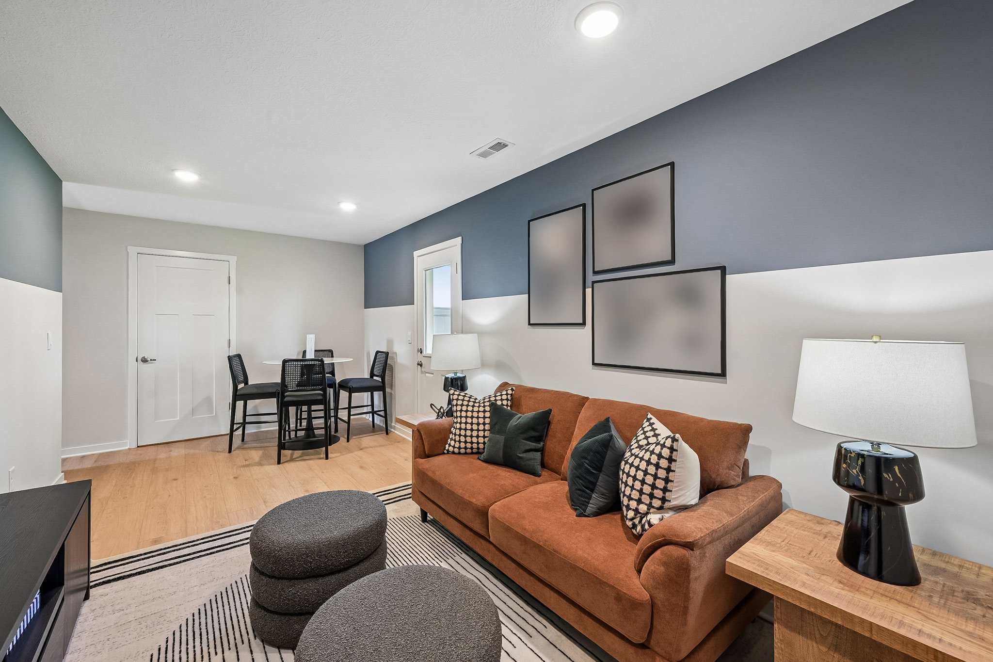 A cozy living room featuring a brown sofa with decorative pillows, a wooden side table, a round ottoman, a dining table set for two, and wall art in neutral tones.