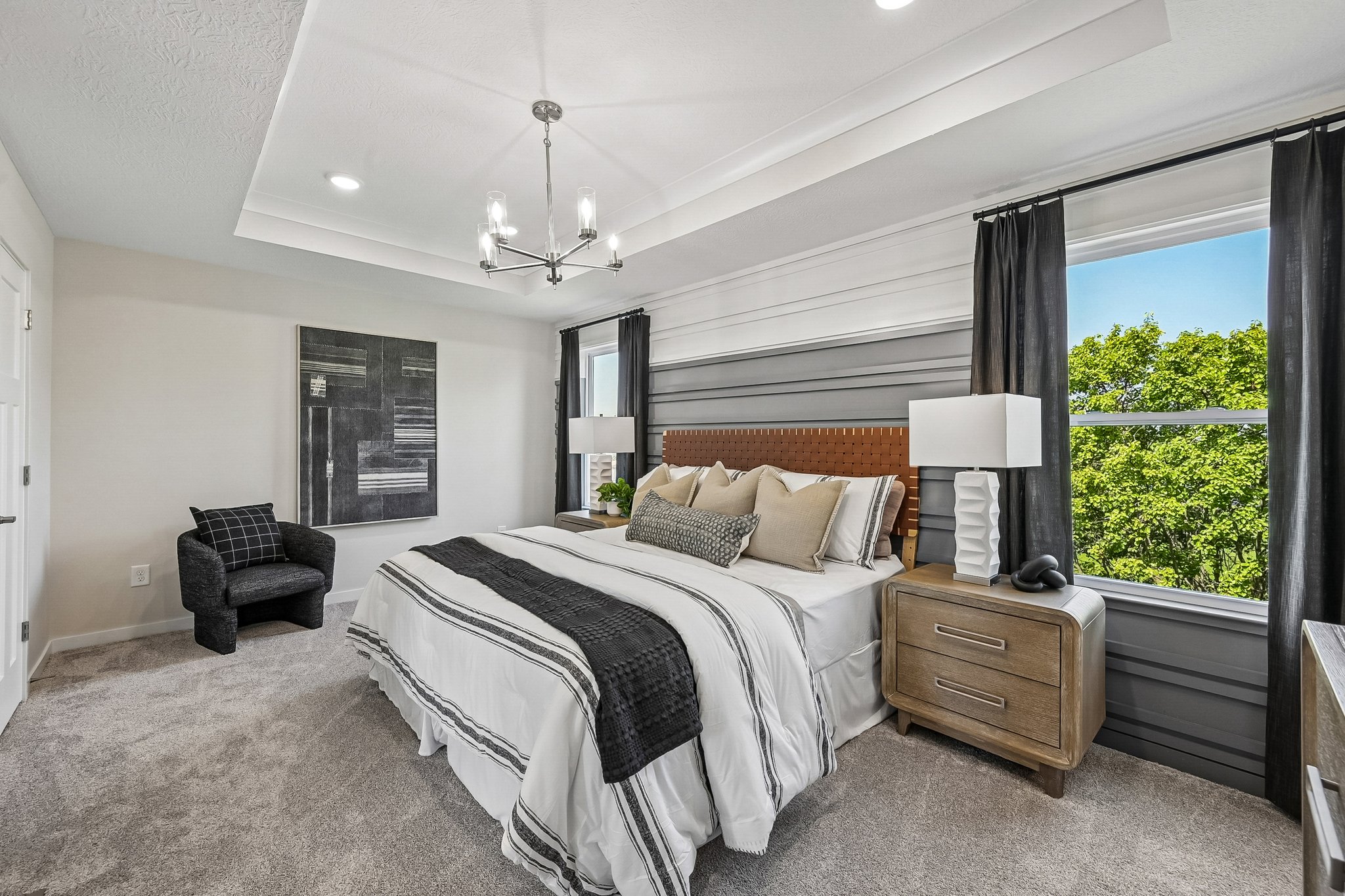A modern bedroom featuring a queen-sized bed with decorative pillows, a nightstand with a lamp, a small chair, and a window with greenery visible outside.