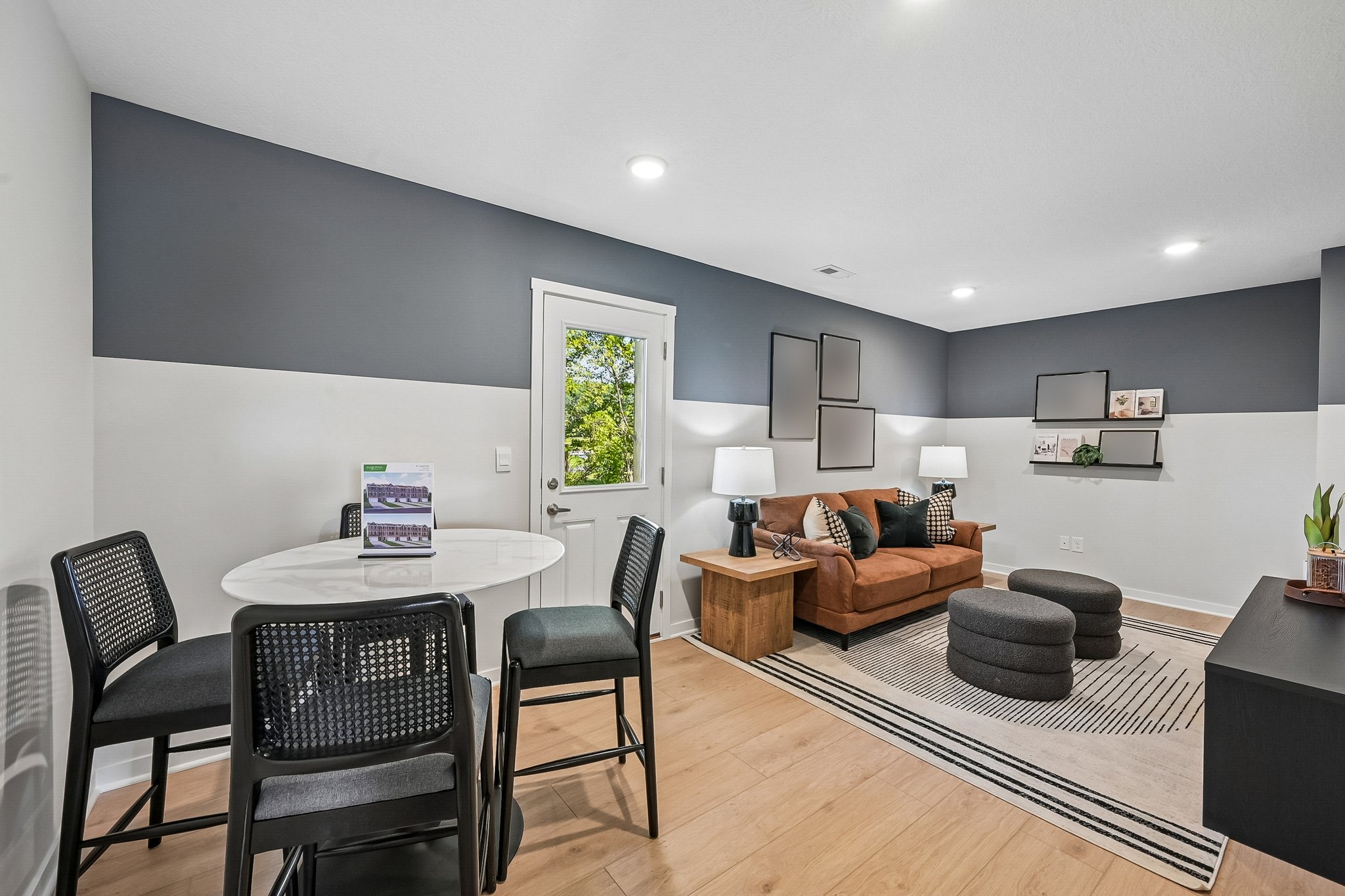 A modern living area featuring a brown couch, round ottoman, and a dining table with black chairs, beside a sliding glass door that provides natural light.