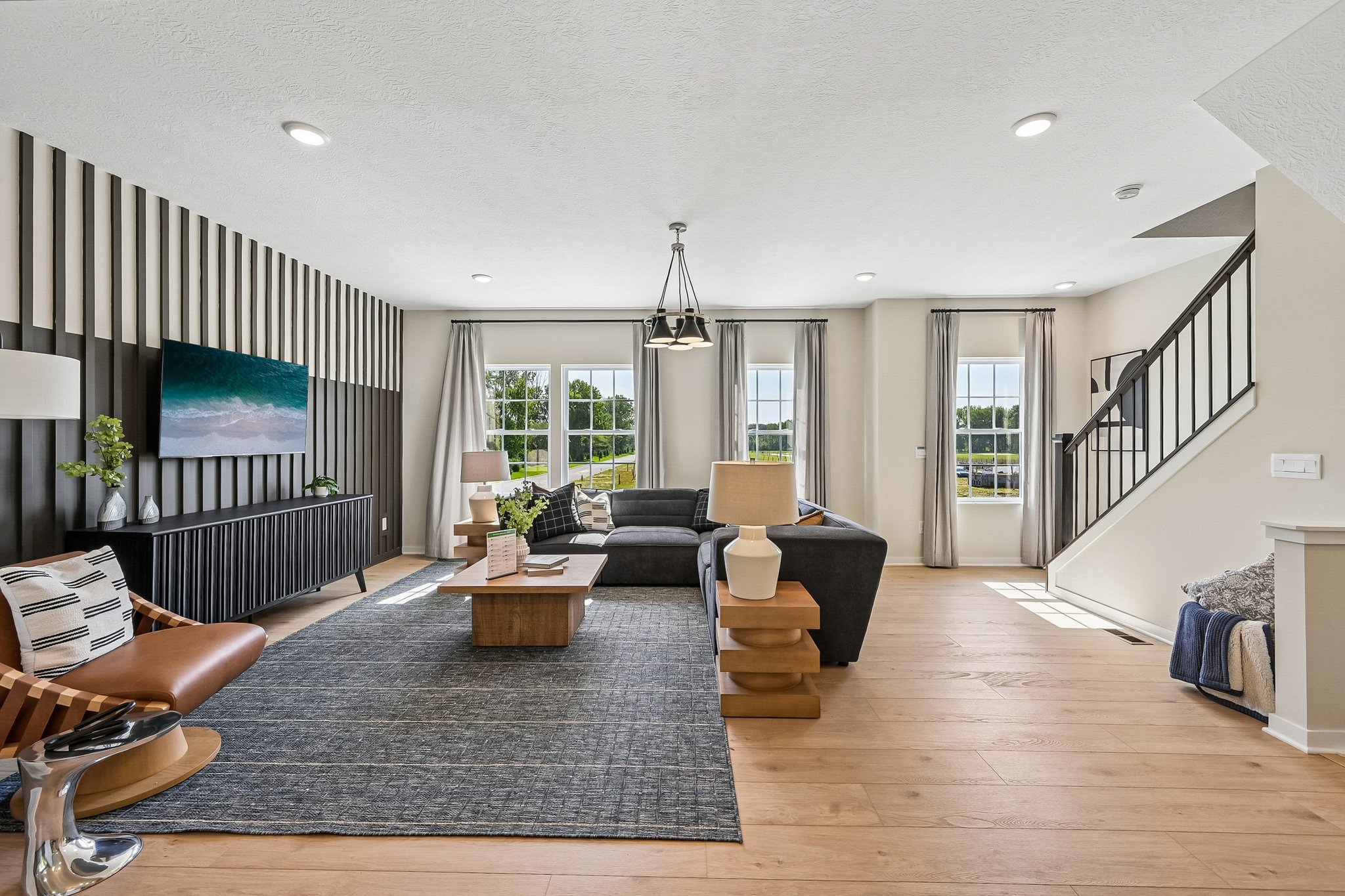 Modern living room with a sectional sofa, wood coffee table, and large windows, featuring a staircase and decorative wall panels.
