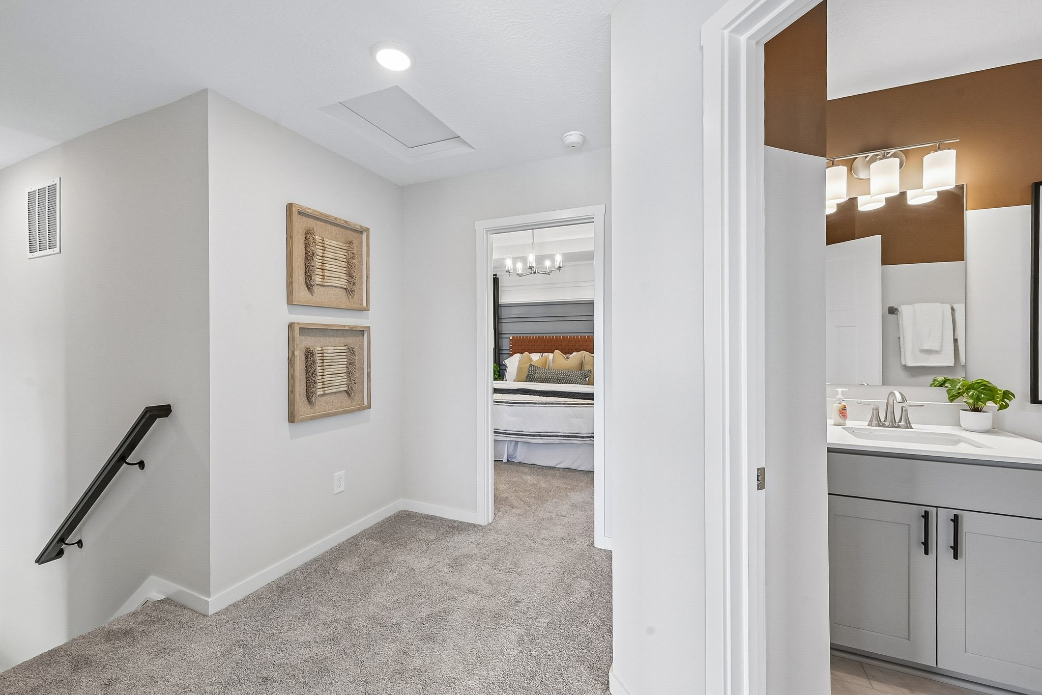 Brightly lit hallway with beige carpet, framed artwork on the walls, and a bathroom sink visible in the foreground.