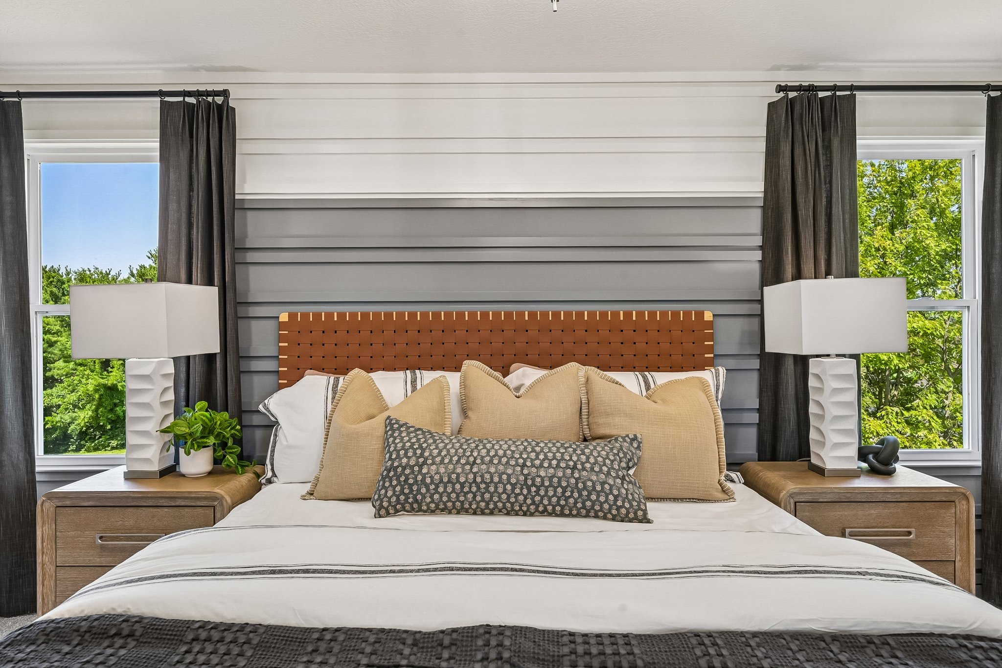 A modern bedroom featuring a neatly made bed with decorative pillows, two bedside tables with lamps, and large windows with sheer curtains.