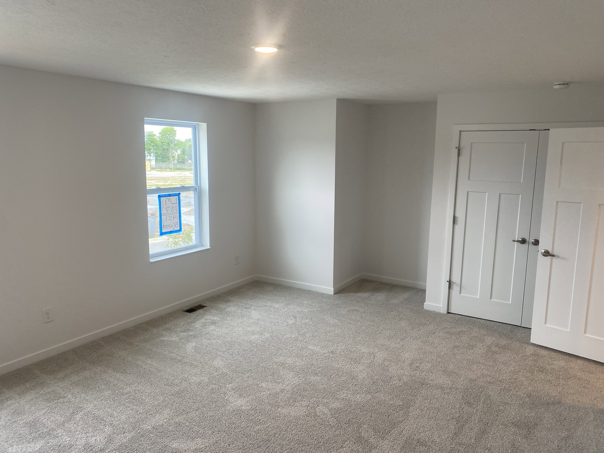 A bright, empty room with light gray carpet, a large window, and a white door leading to another space.