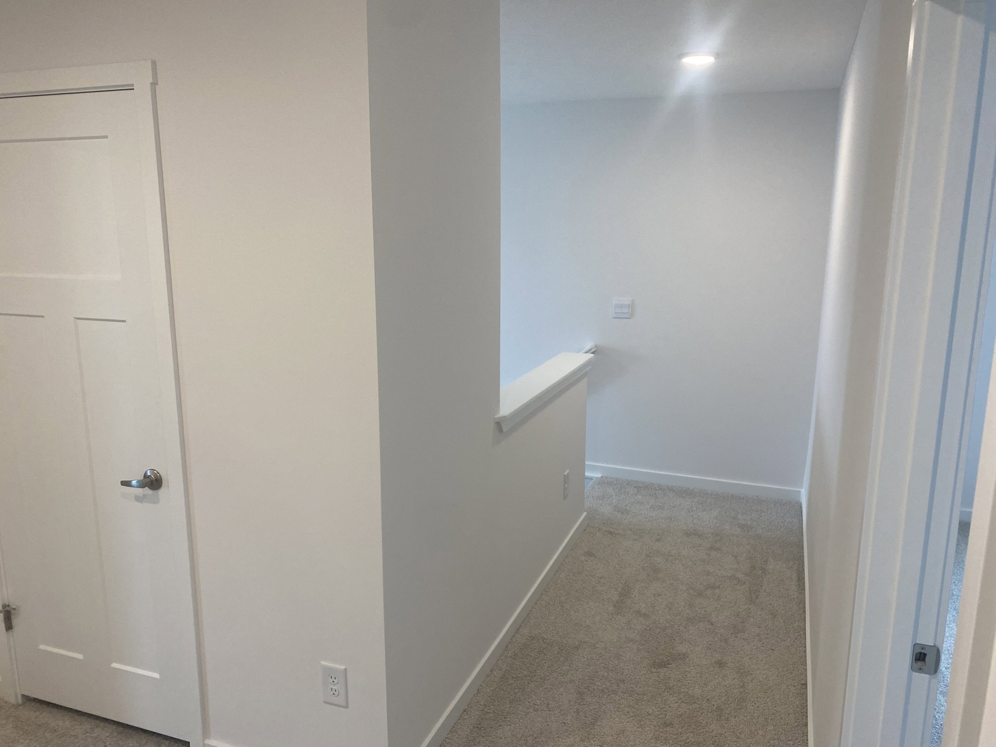 A well-lit hallway with light gray walls and carpet, featuring a closed door on the left and an open doorway on the right.