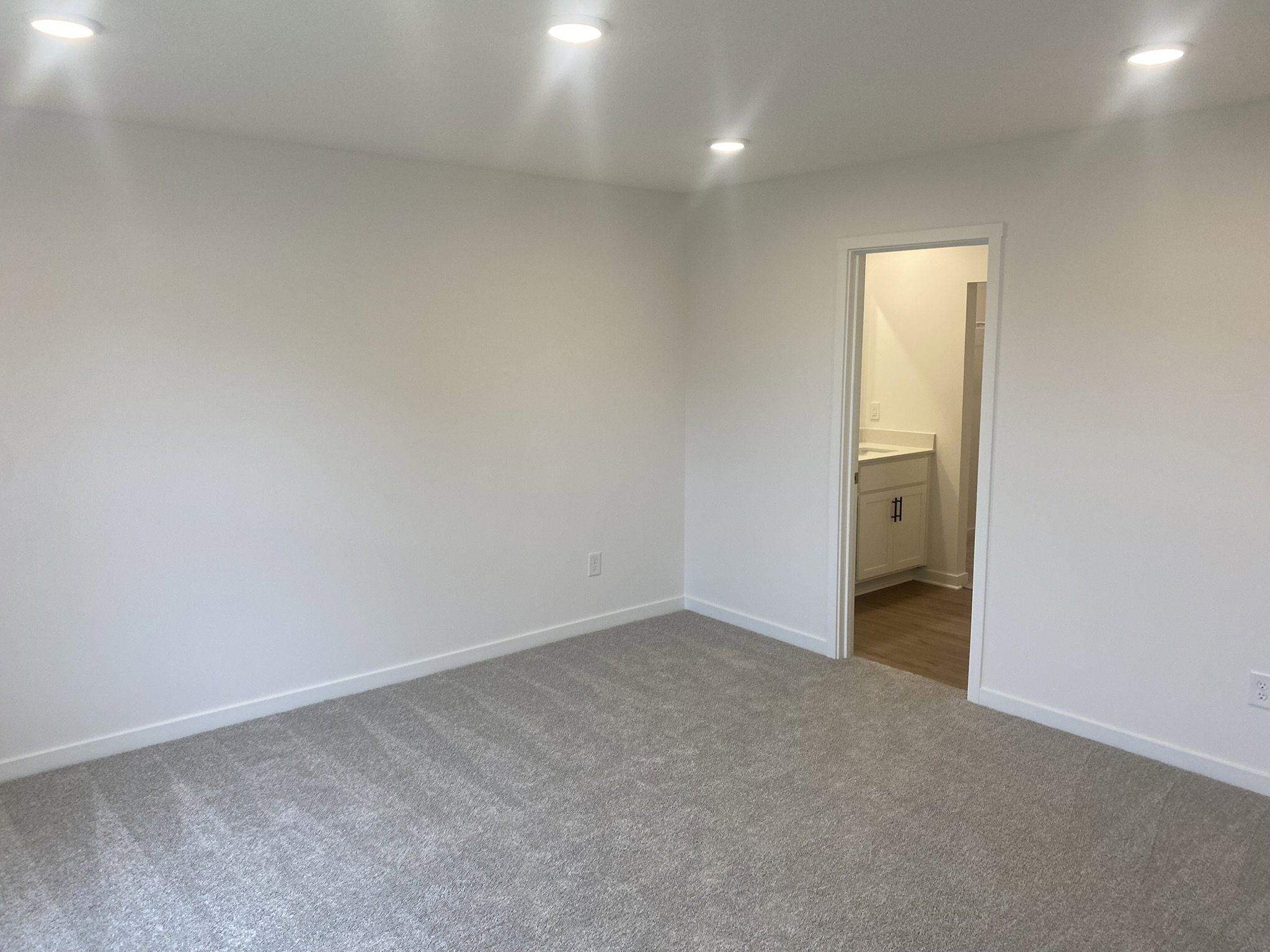 Empty room with light-colored walls, carpeted floor, and an open doorway leading to a bathroom.