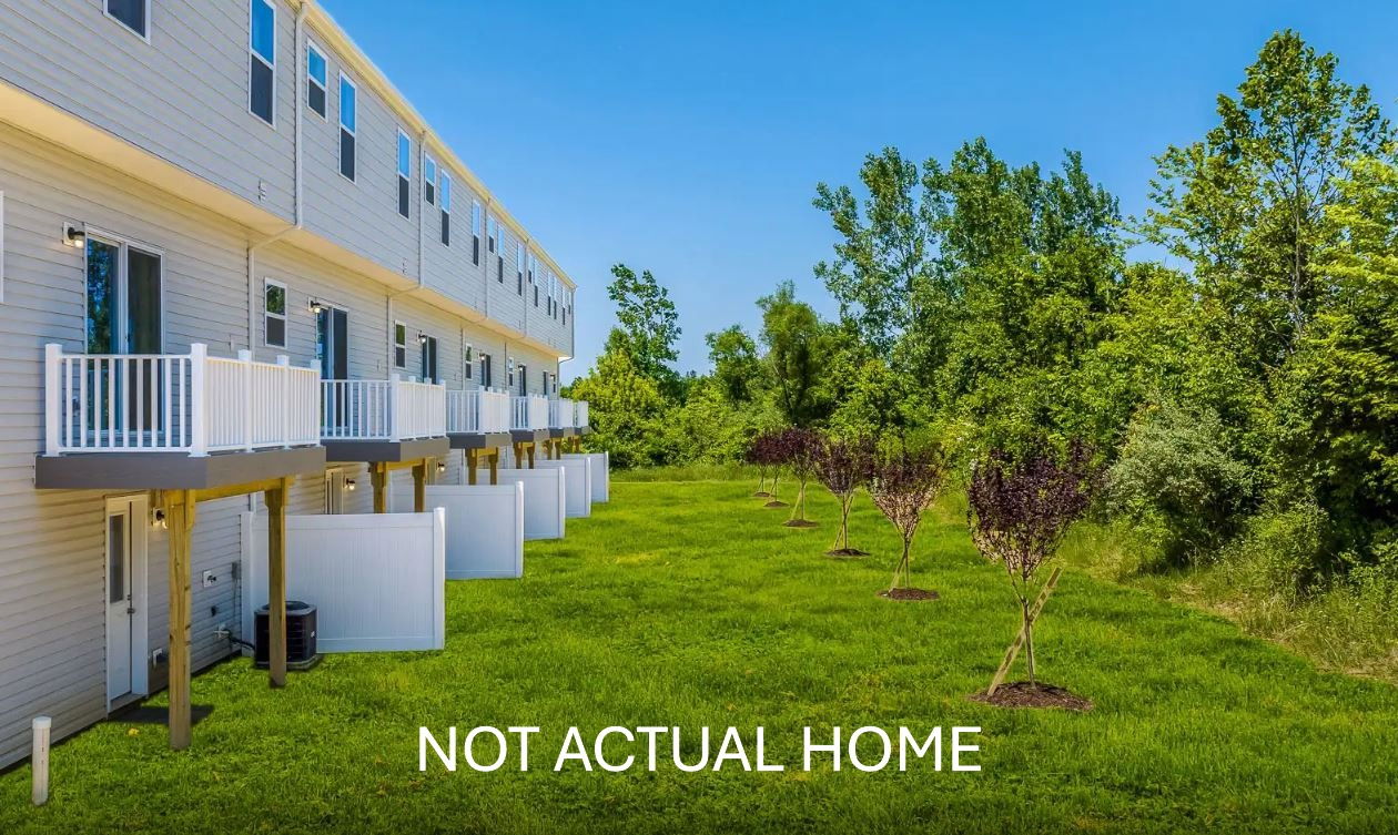 A row of modern townhouses with balconies overlooking a lush green backyard and trees under a bright blue sky.