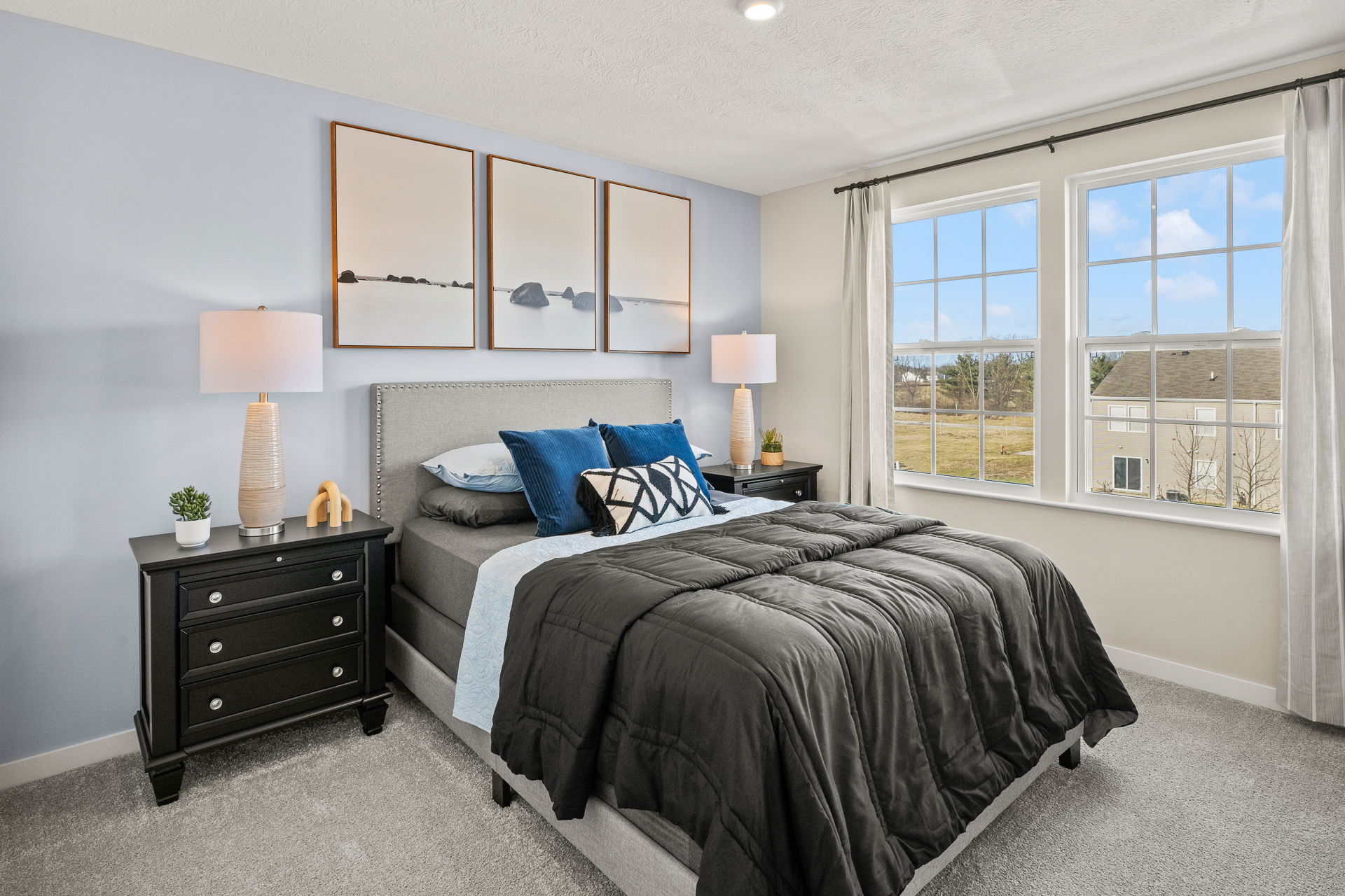 Modern bedroom featuring a gray bed with decorative pillows, stylish nightstands with lamps, and large windows showcasing an outdoor view.