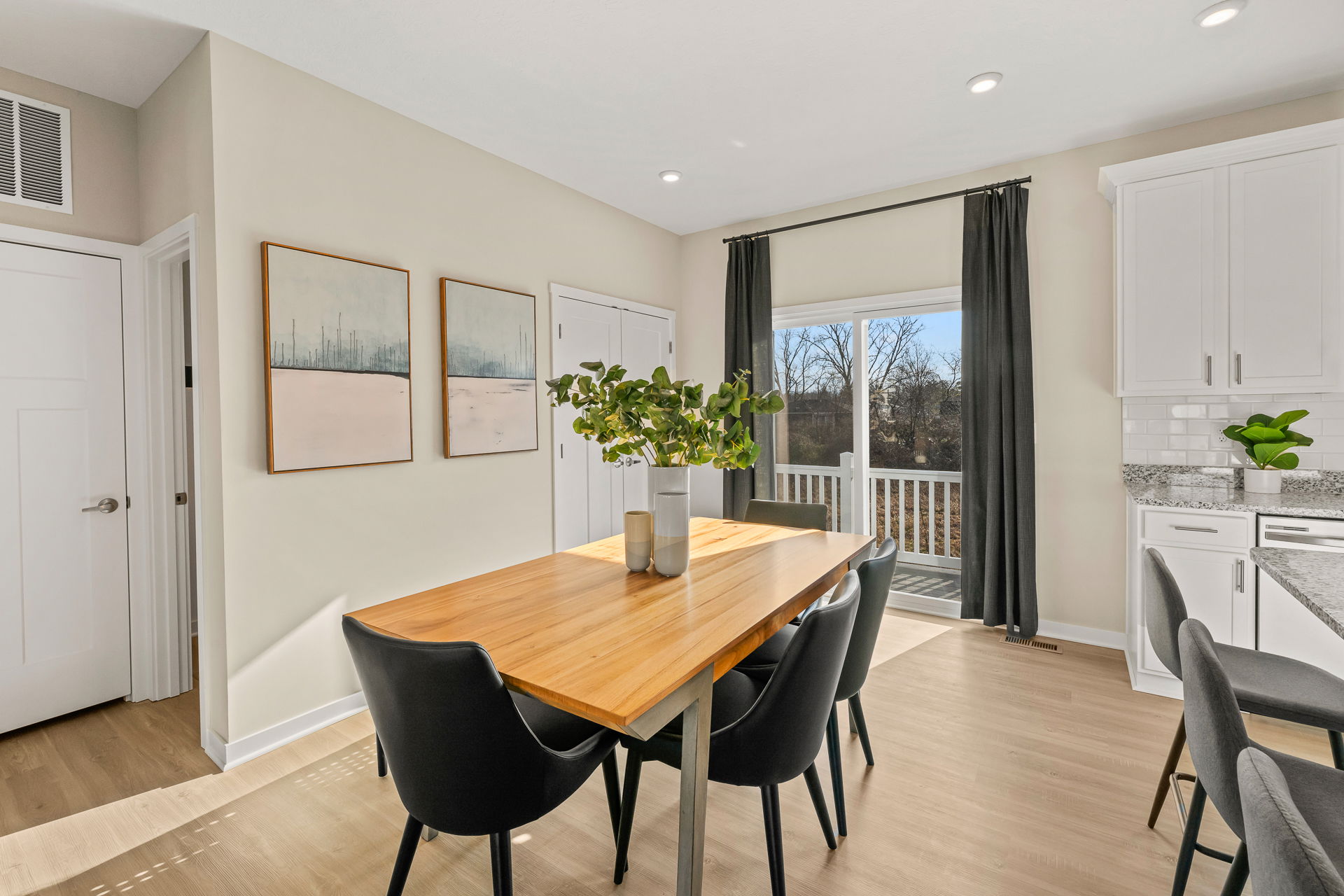 Bright and modern dining area featuring a wooden table, black chairs, and contemporary art on the walls with natural light streaming in from a nearby balcony door.