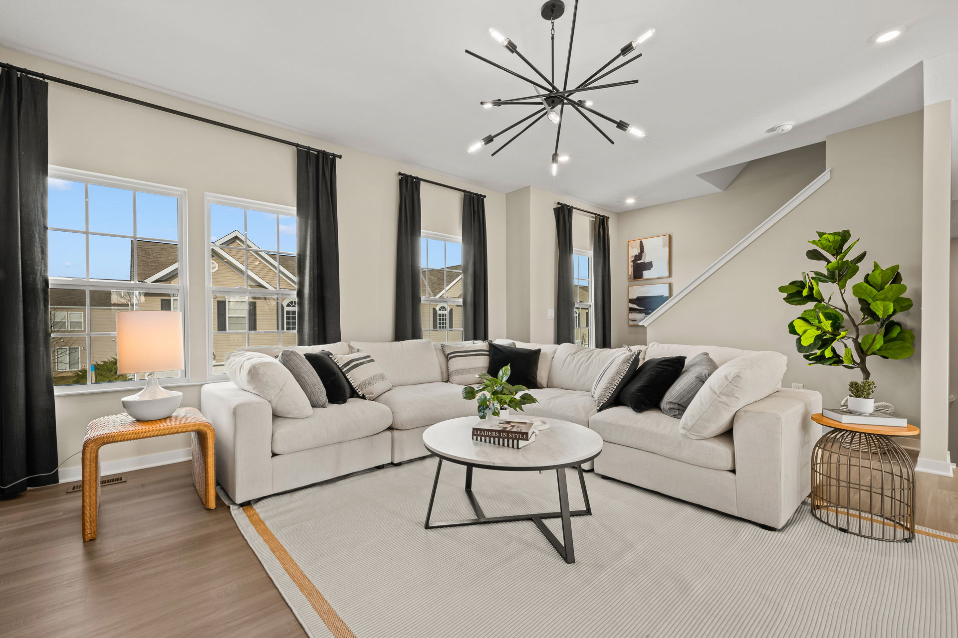 Modern living room featuring a comfortable beige sectional sofa with decorative pillows, a stylish coffee table, and large windows with black curtains allowing natural light.