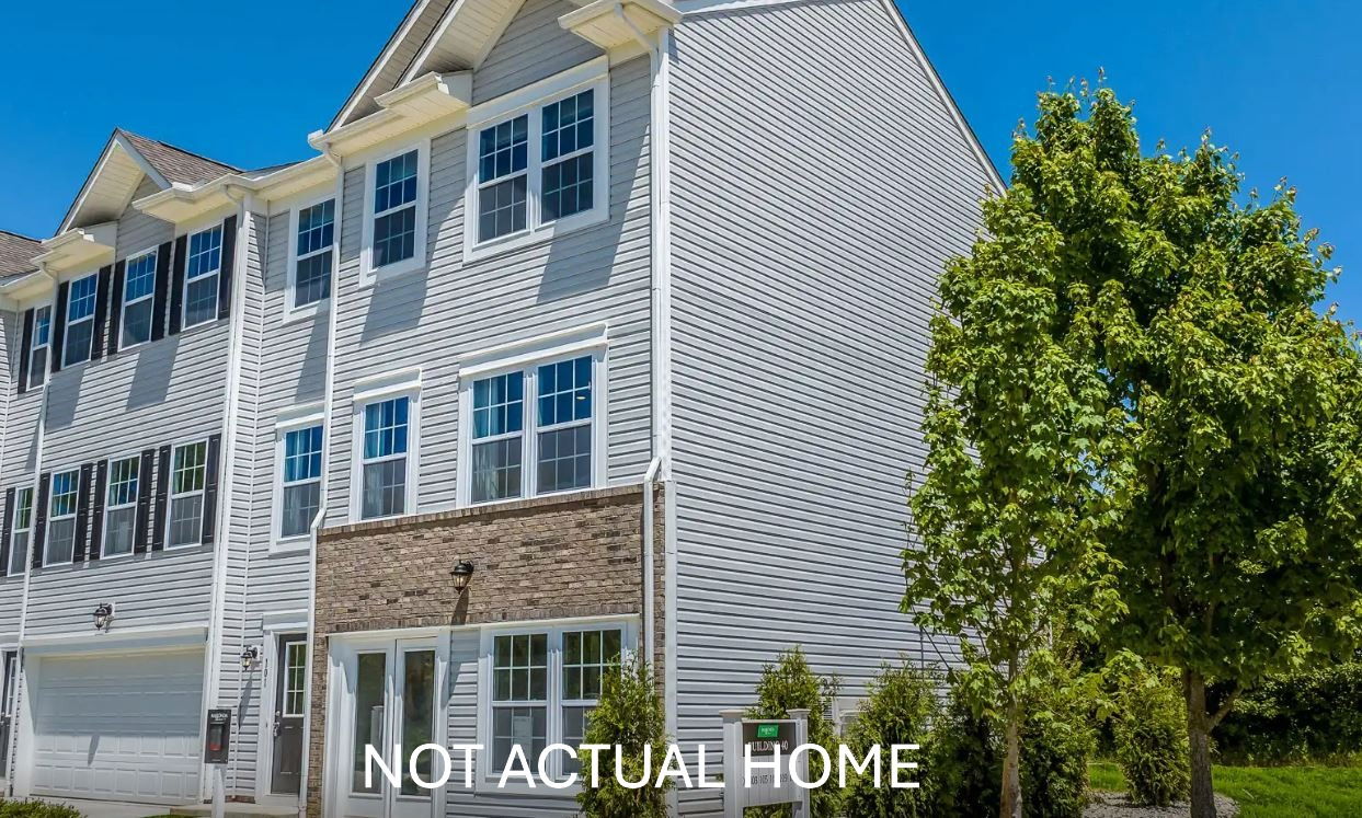 A modern three-story townhouse with gray siding, large windows, and a brick accent, surrounded by green trees under a clear blue sky.