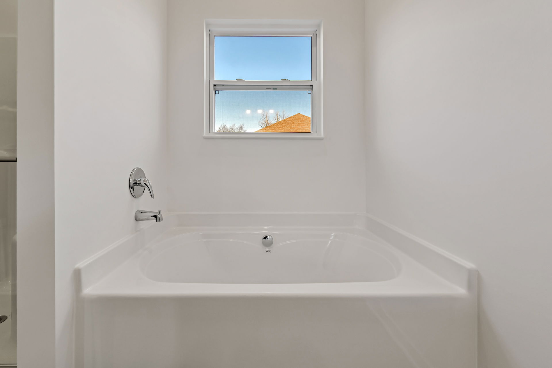Modern white bathtub with chrome fixtures situated below a window in a minimalist bathroom.