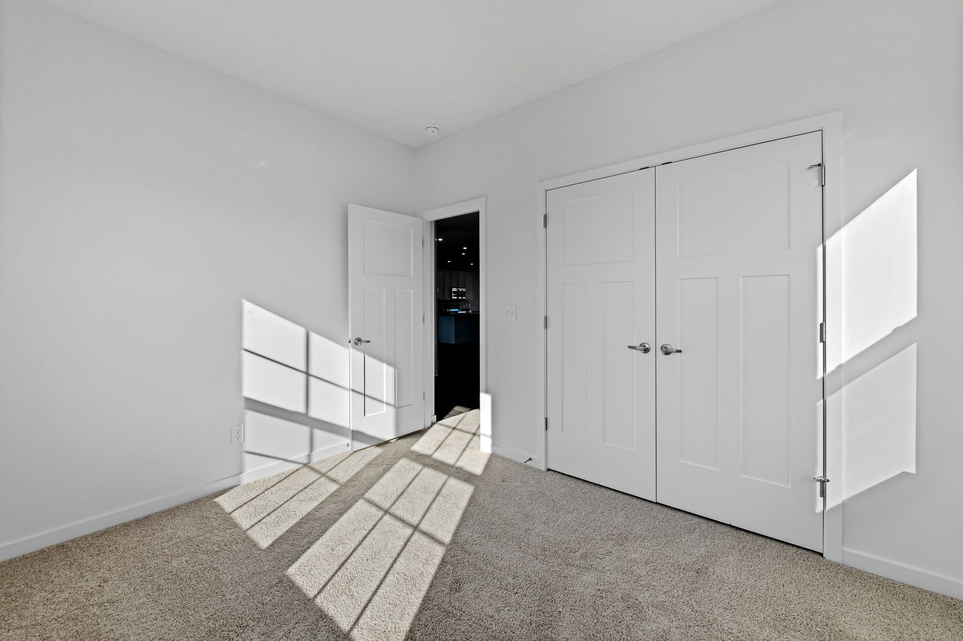 Empty bright room with white walls and closed double doors, featuring sunlight streaming through a window onto beige carpet flooring.