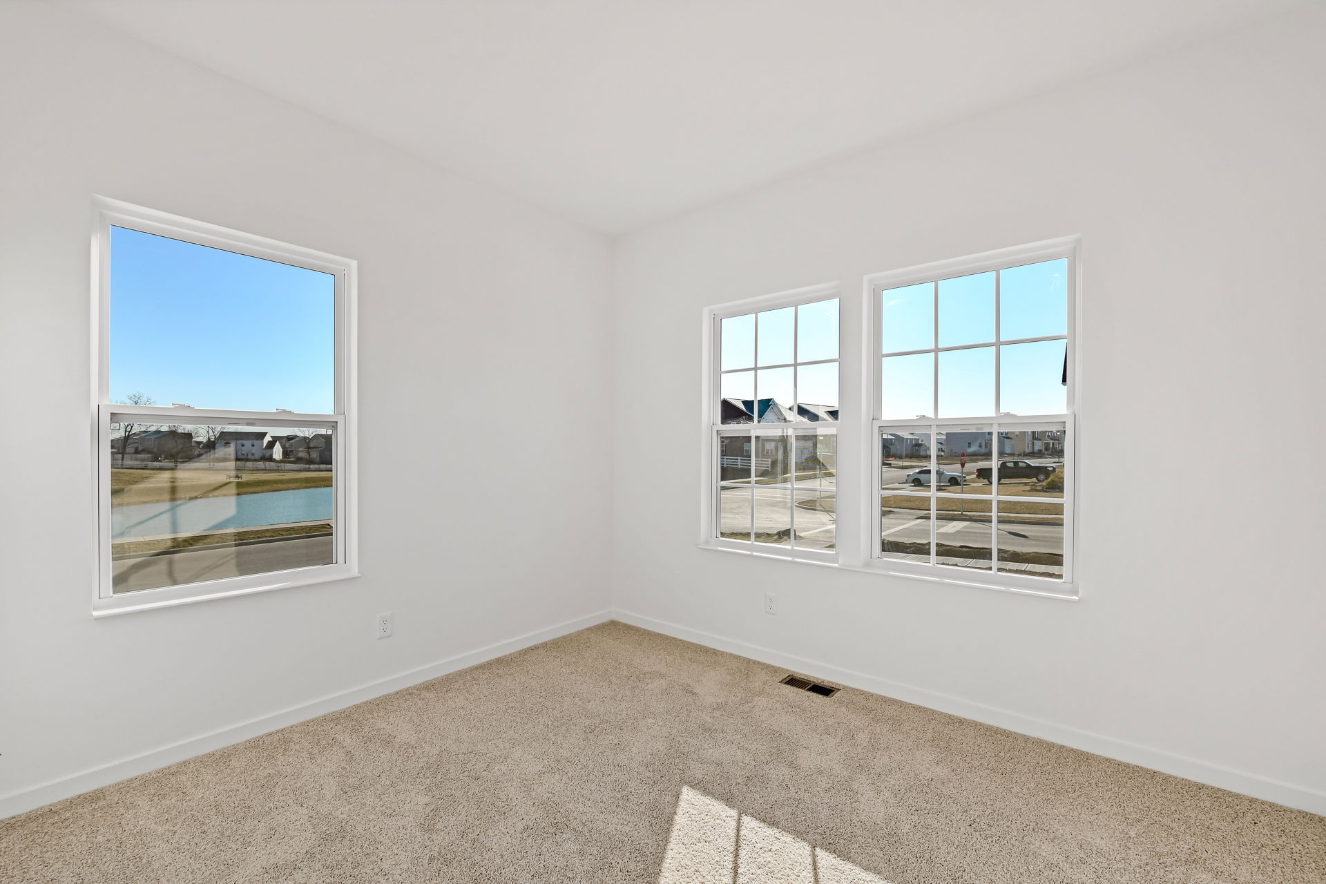 Bright, empty room with beige carpet and large windows overlooking a suburban neighborhood and pond on a sunny day.