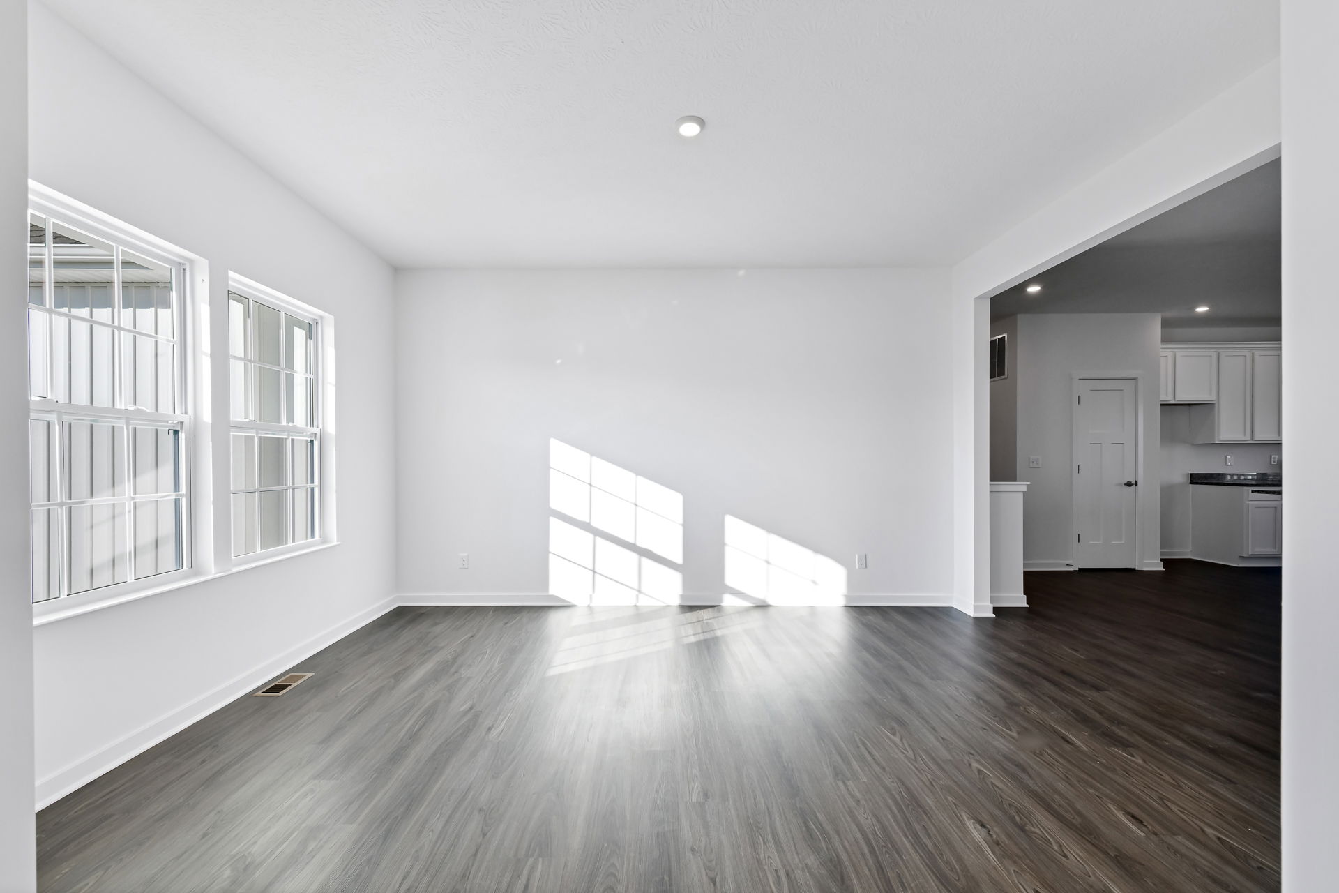 Bright, empty room with large windows and sunlight on hardwood floors, leading to an adjacent kitchen area.