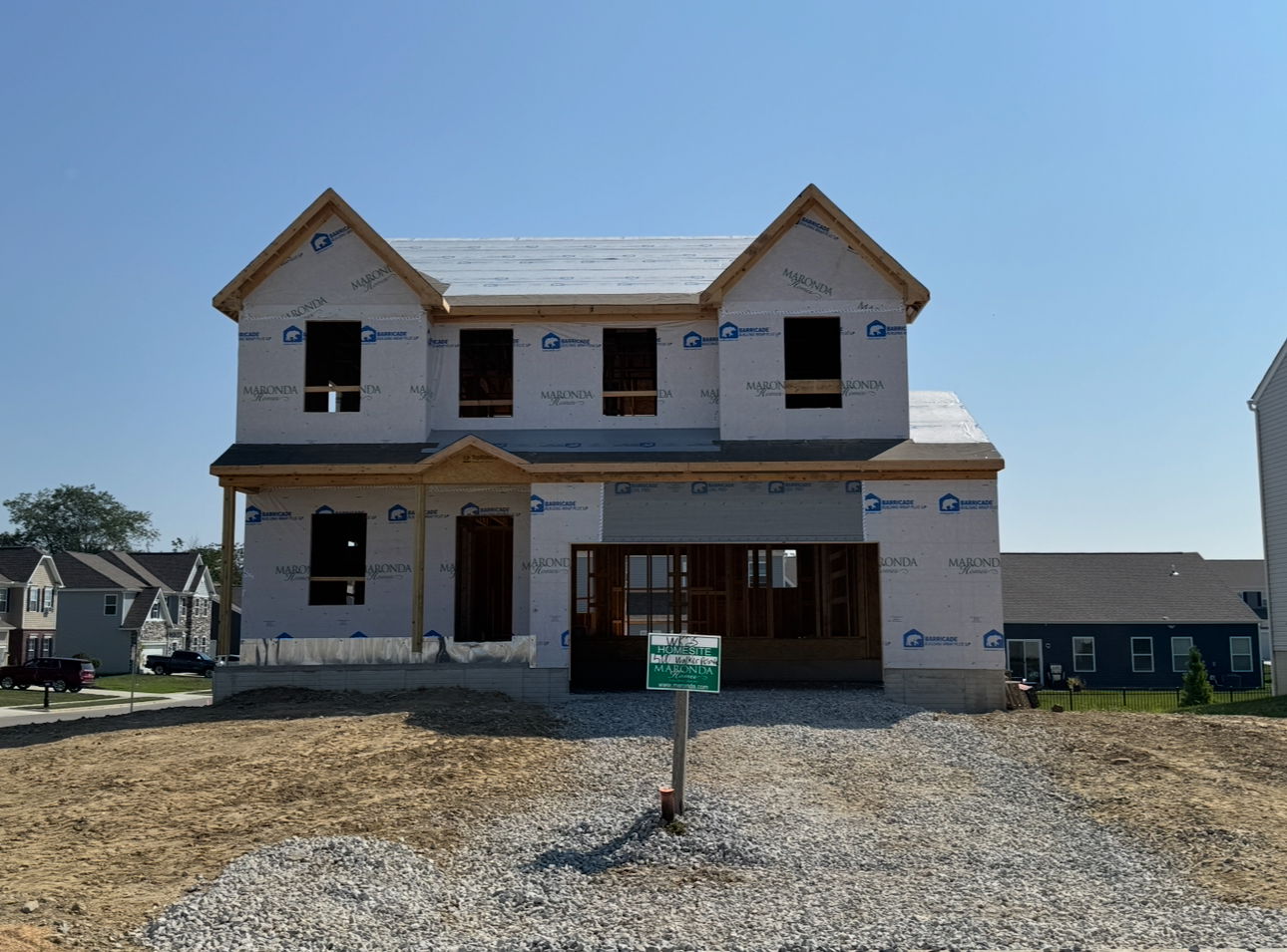 Two-story house under construction with visible sheathing and a gravel driveway in a developing neighborhood.
